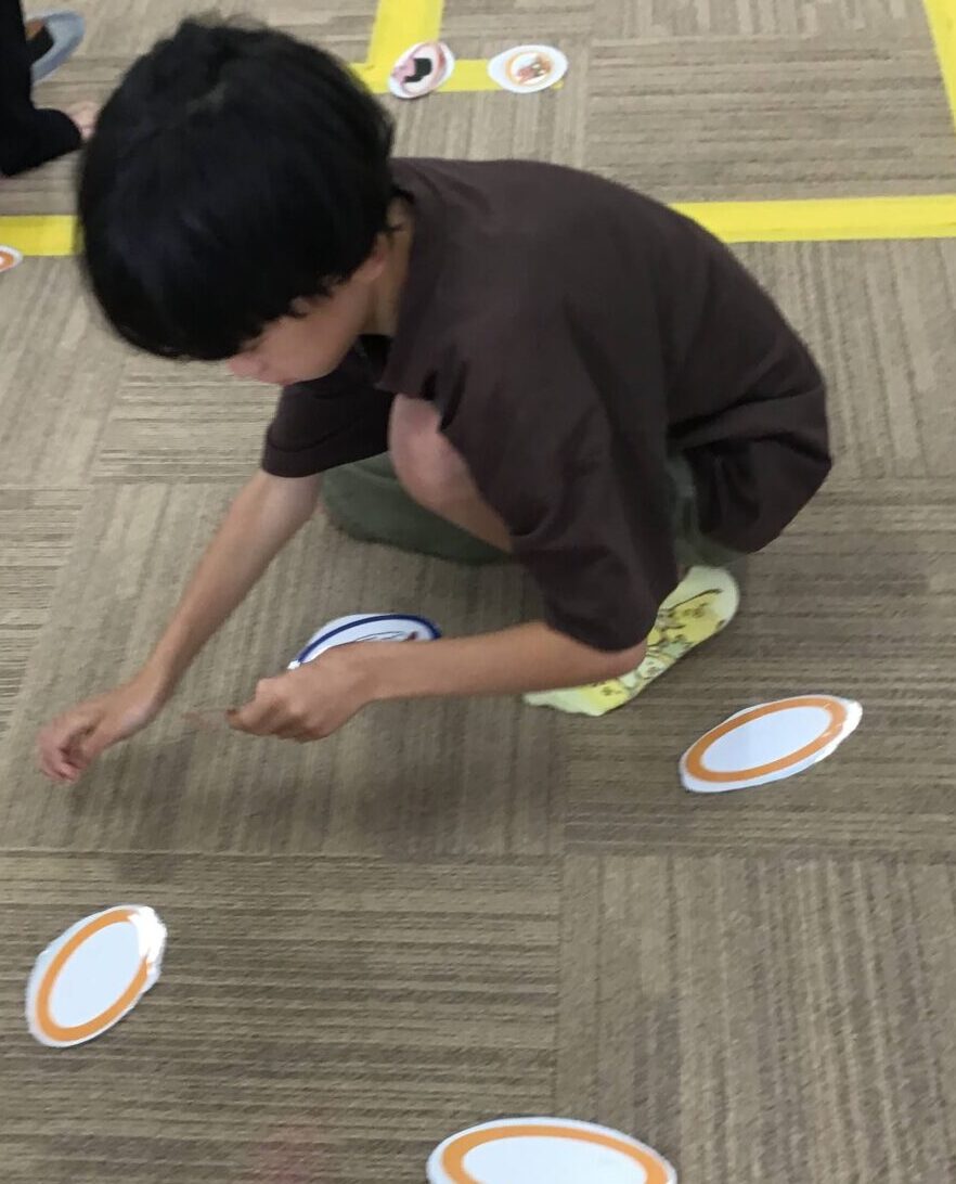 Child crouching on carpet picking up orange-edged circular cards scattered on the floor for a game/activity.