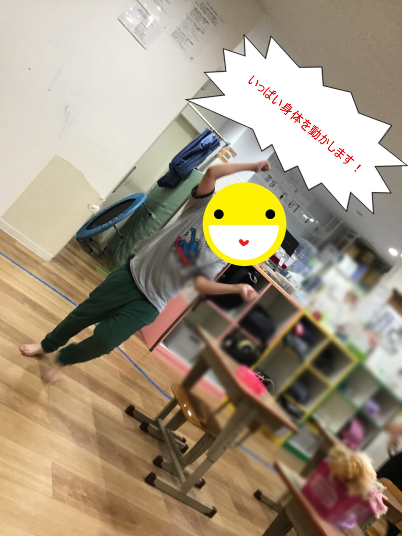 Child standing in a classroom or gym, raising an arm as part of a activity; small trampoline and shelves visible in the background; Japanese burst text says 'いっぱい身体を動かします！' (We move our bodies a lot).