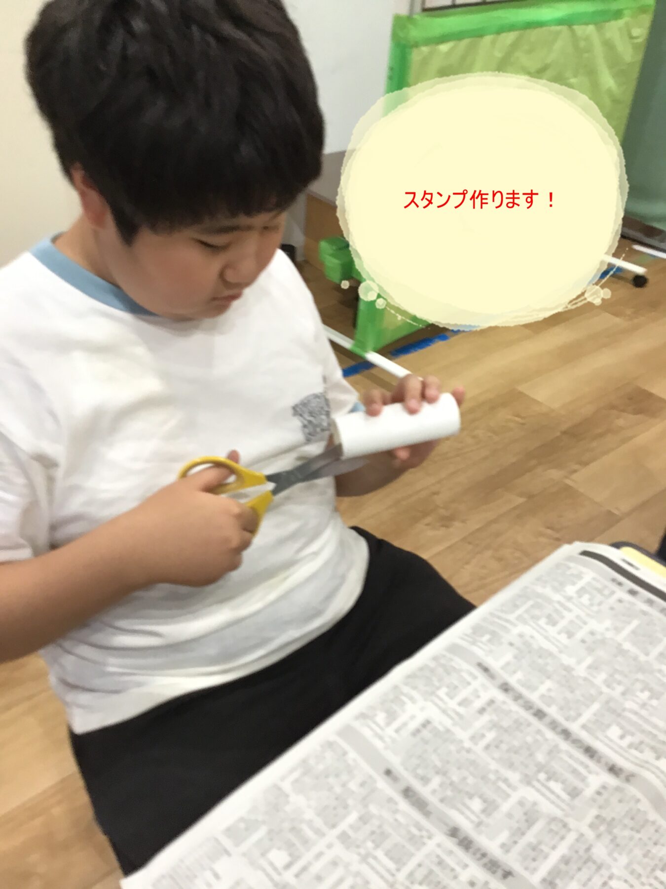 Child sits at a table, cutting a white cardboard tube with yellow scissors for a stamp craft, with a newspaper on the table and a large Japanese text overlay saying “スタンプ作ります！” (We’ll make stamps!).