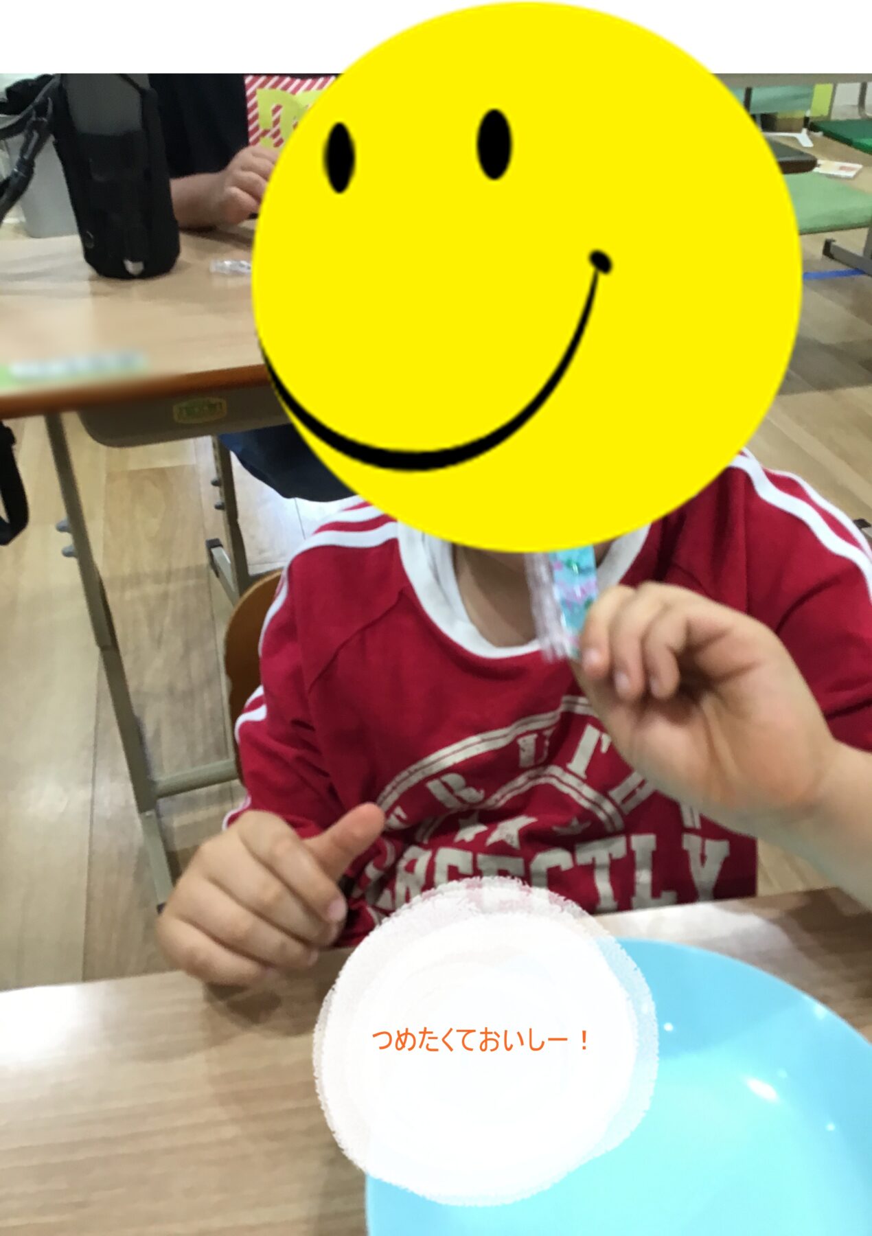 Child in a red shirt sits at a table eating a snack, with a large yellow smiley sticker covering their face for privacy.