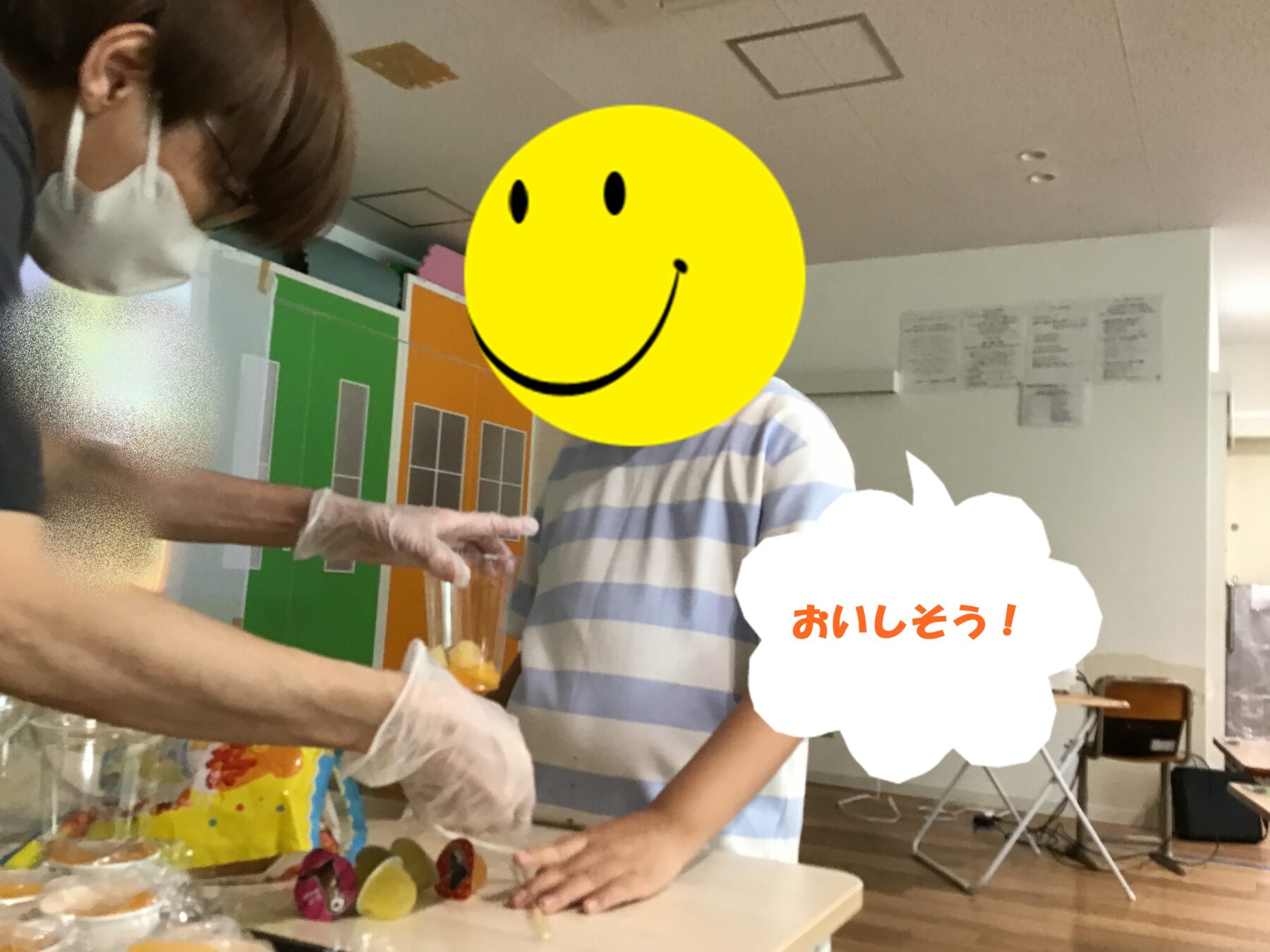 A person wearing a mask and gloves hands a cup of mixed fruit to a child in a bright, classroom-like kitchen. A large yellow smiley face covers another person’s face, and a speech bubble reads in Japanese: “おいしそう!” (looks delicious).
