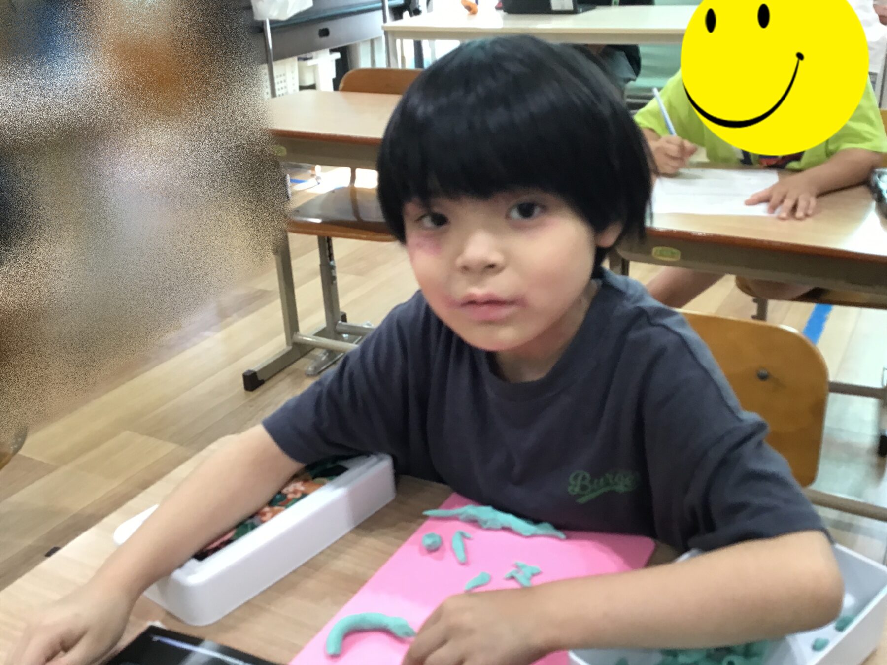 Child with black hair sits at a table, shaping blue-green clay on pink paper during a classroom craft activity; other student with a yellow smiley face sits in back.