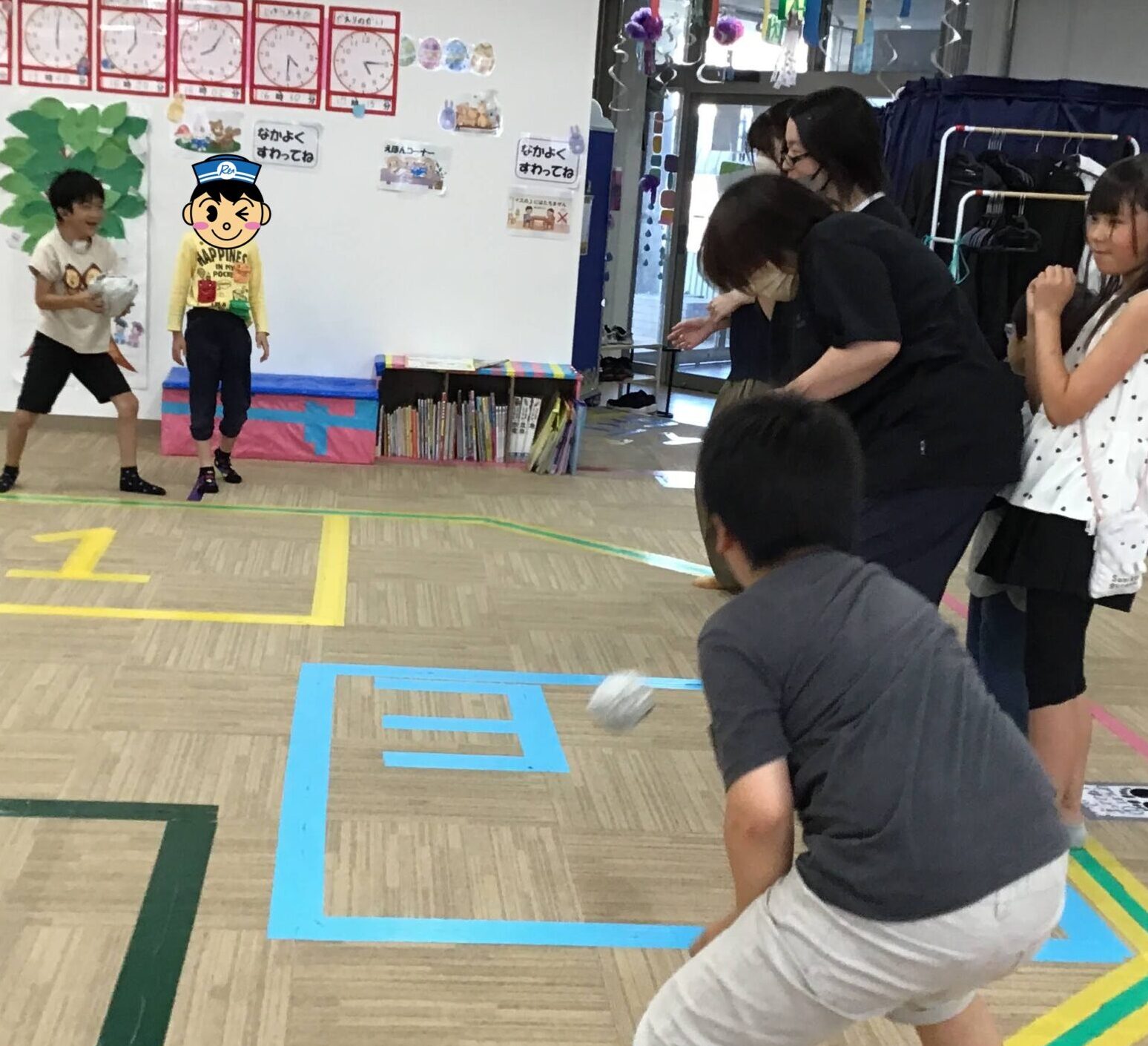 Two children at left throw a soft object toward a group of people watching; a gym-like room with colorful floor markings and posters on the wall behind them.