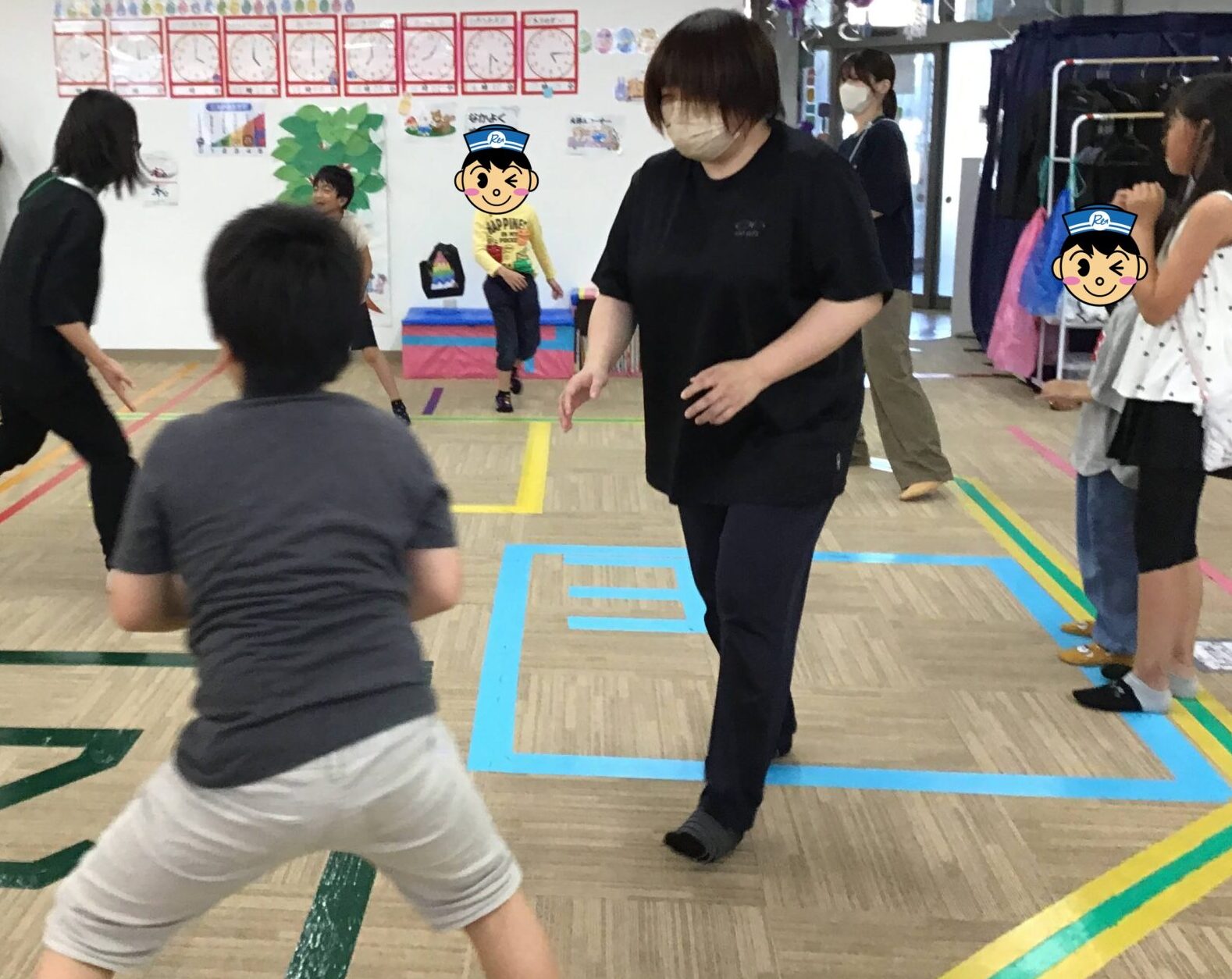 People playing a group game in a colorful indoor gym; kids run while adults watch, wearing masks, floor markings guide activity.