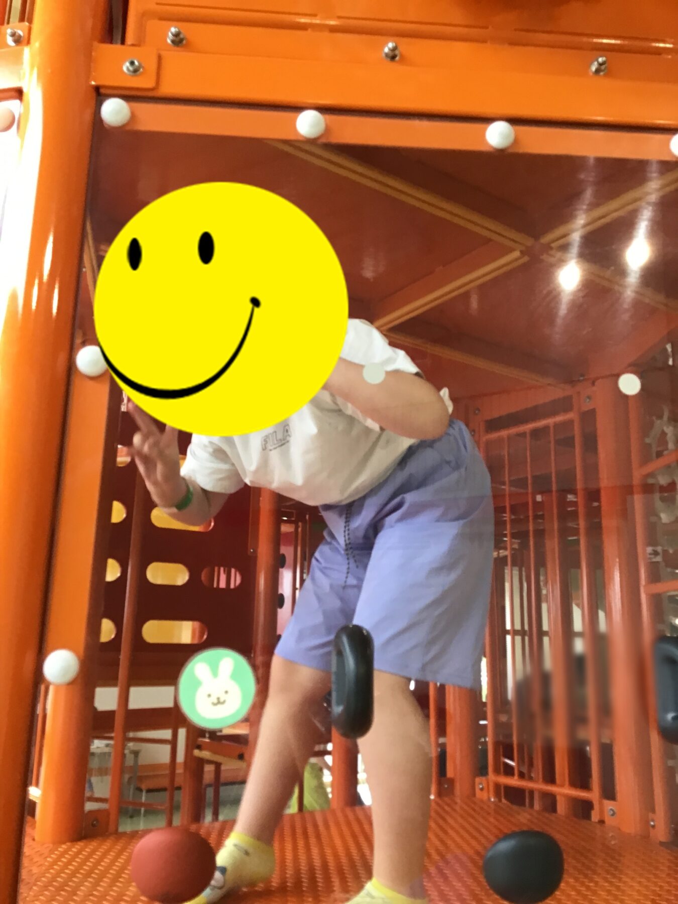 Child climbing inside an orange indoor play structure, wearing a white shirt and light blue shorts, with colorful balls on the orange floor.