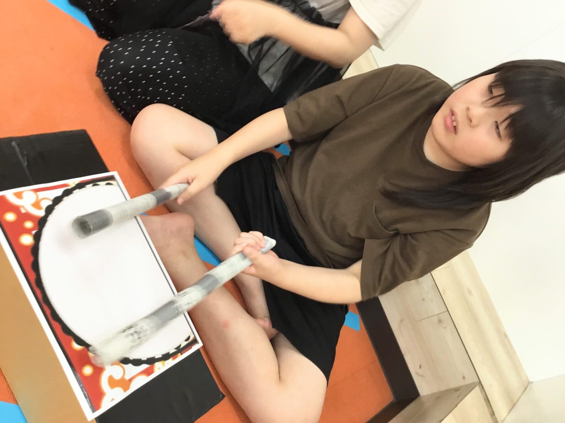 Child in a brown shirt sits on an orange mat, holding two white cylindrical sticks near a framed poster.] ,