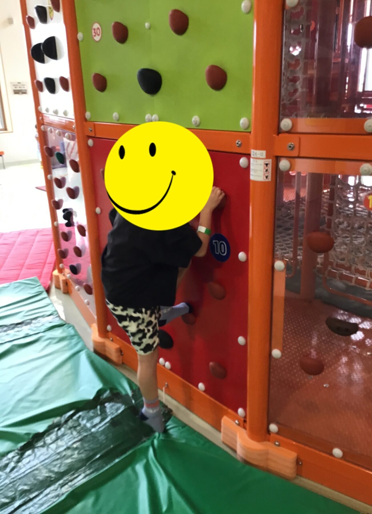 Child wearing black shirt and patterned shorts climbs an indoor orange climbing wall with holds, padded green floor beneath.
