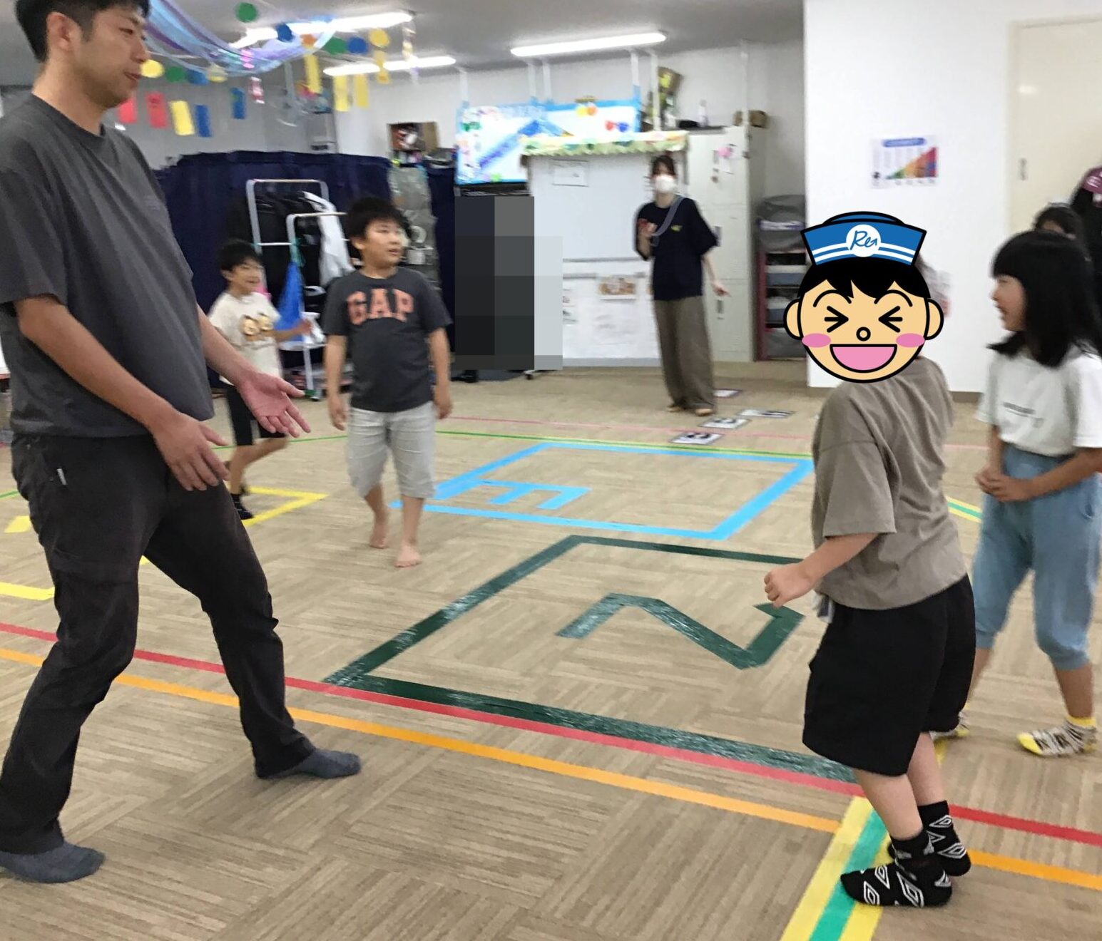 Adult instructor in a dark gray shirt leads two children in an indoor activity, standing on a gym floor with colorful taped lines.