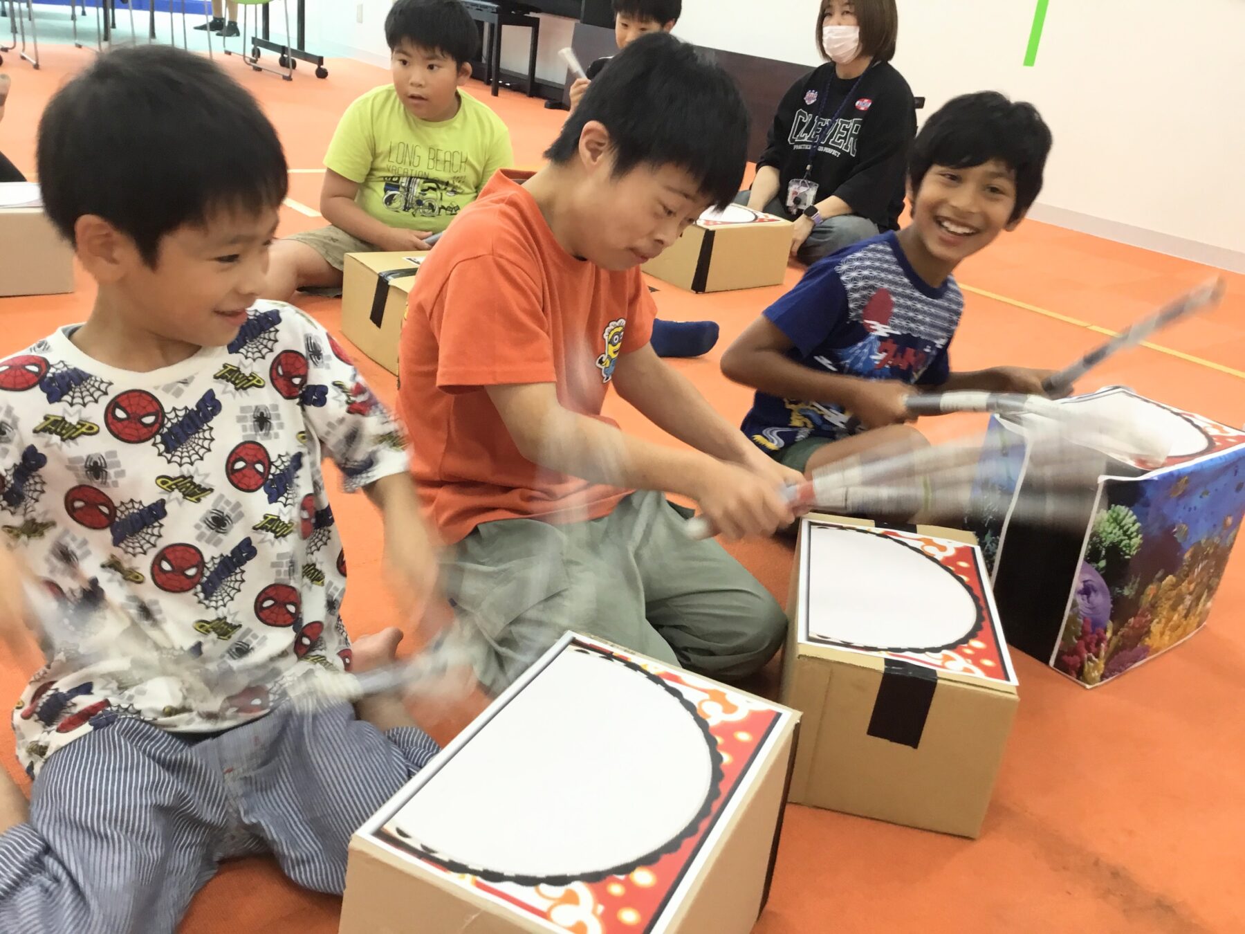 Children sit on an orange gym floor, using cardboard box drums and drumsticks during a group activity, with a masked adult supervising in the background.