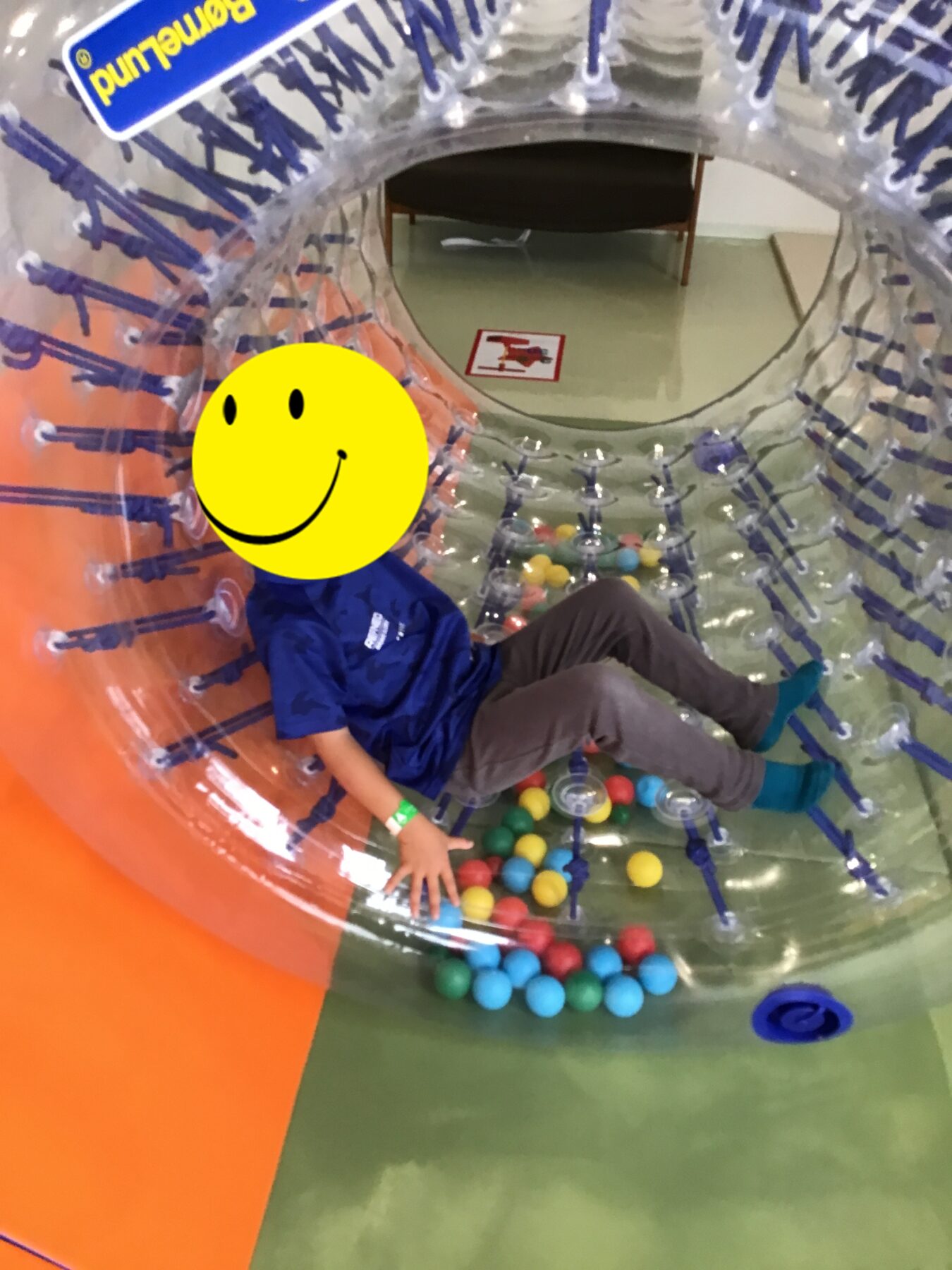 Child in a blue shirt sits inside a clear inflatable play tunnel filled with colorful plastic balls in an indoor play area.