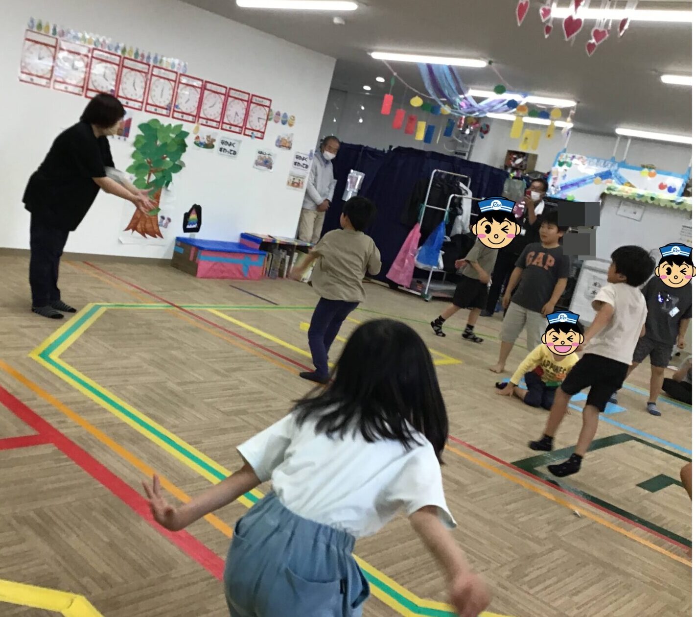 Teacher leads a kids’ running activity in a school gym, with children weaving around colorful taped lines on the floor and wall decorations in the background (tree cutout and artwork).