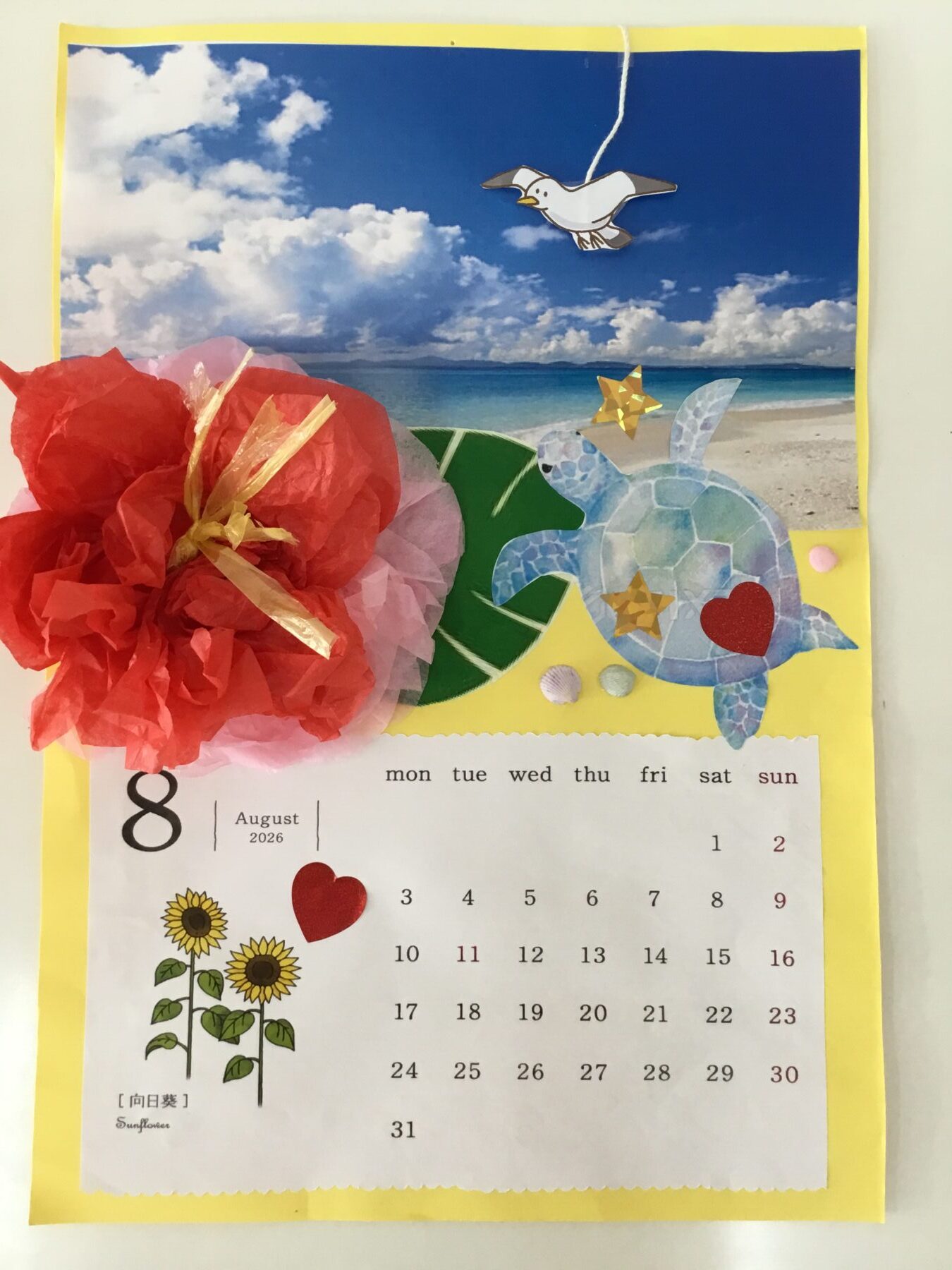 Decorative collage calendar page with a mosaic blue sea turtle, red heart, tropical flowers, and tissue paper decorations against a yellow border.