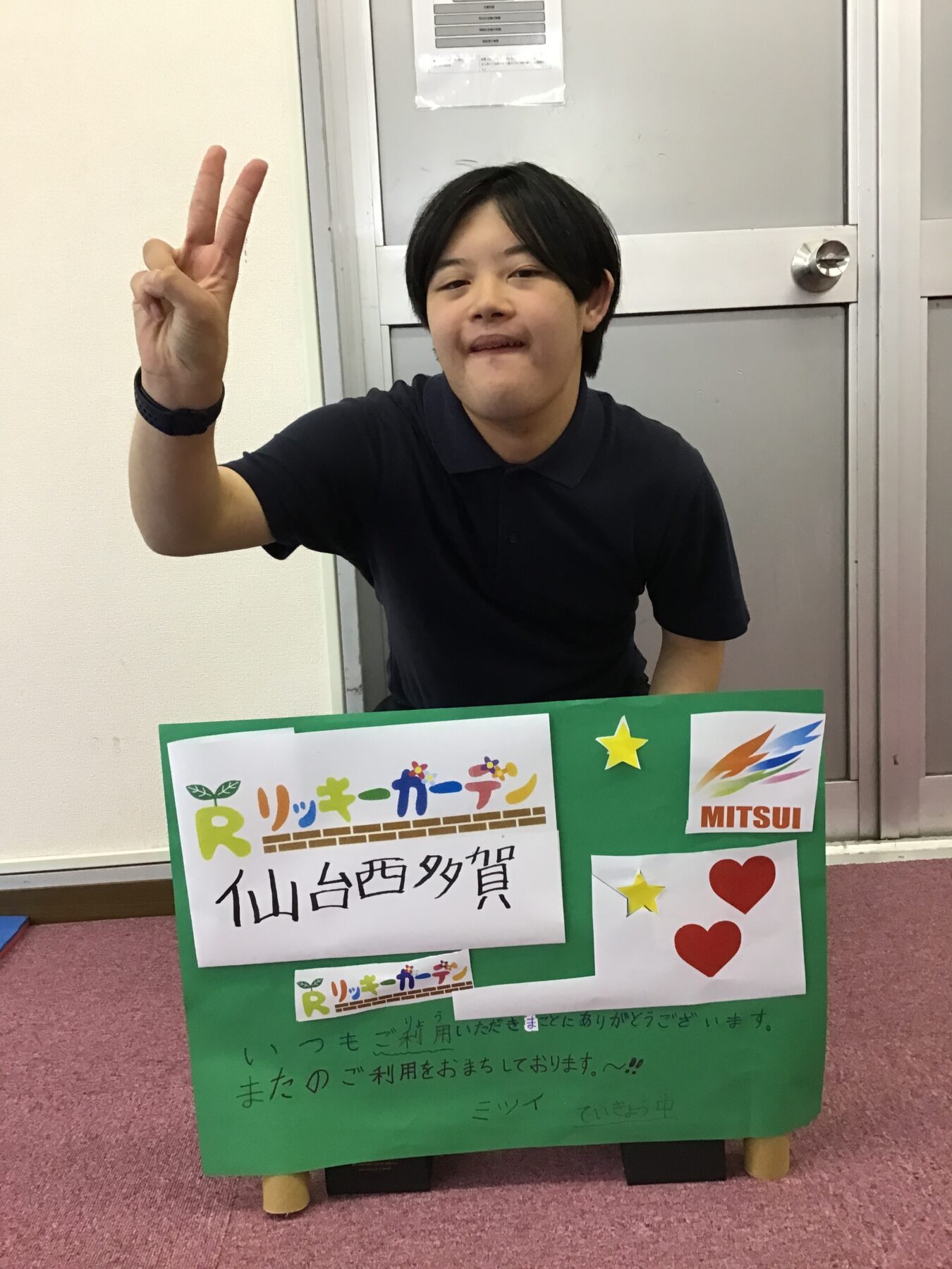 Young man in a black polo shirt sits indoors, making a peace sign; a green poster with logos and Japanese text is in front.
