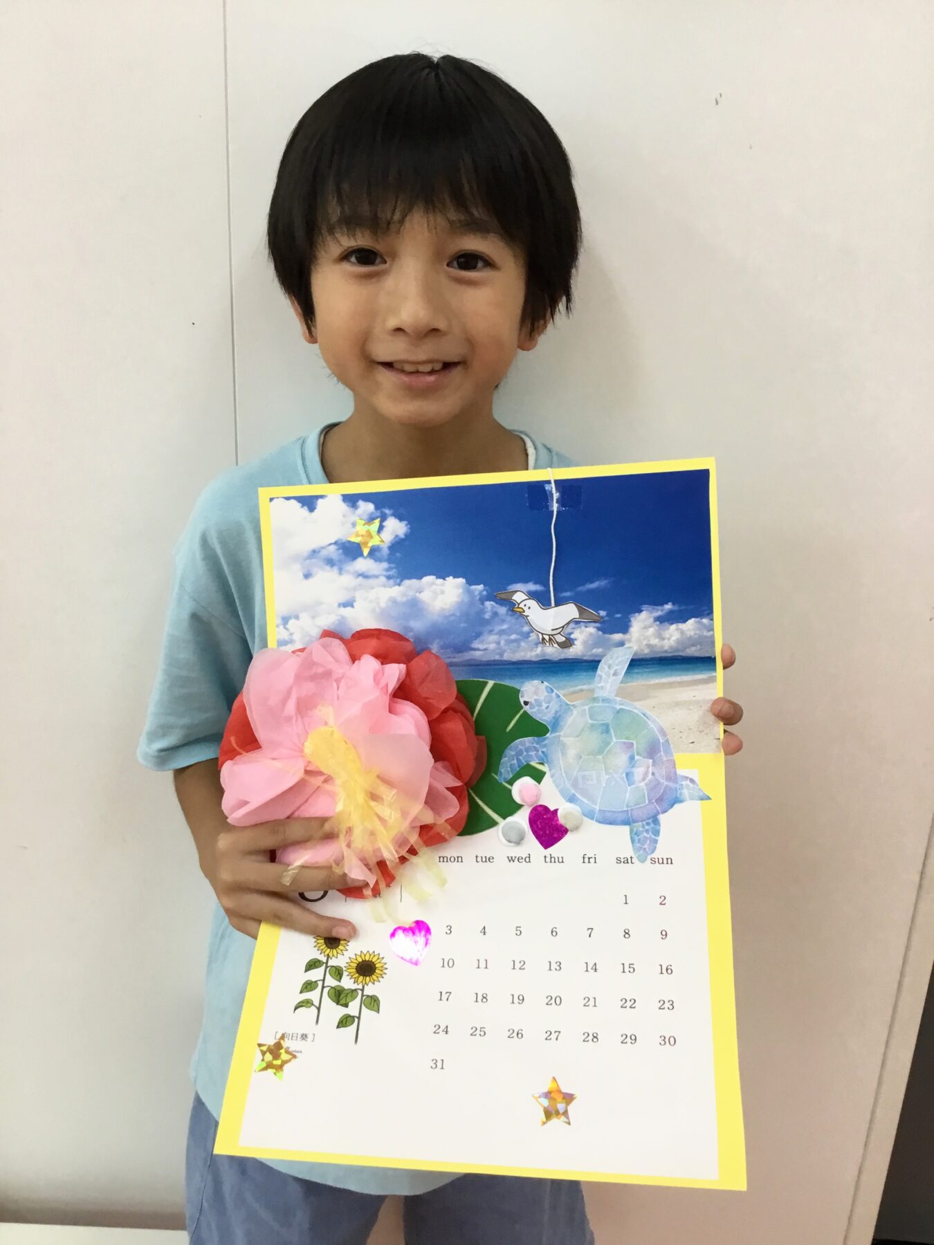 Young boy smiling at the camera, wearing a light blue shirt and holding a bright, decorated calendar with a large pink paper flower attached to it.