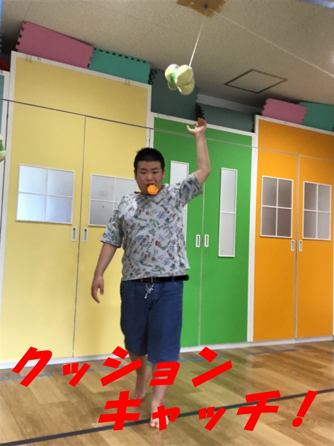 Person standing indoors on a wooden floor, one arm raised, wearing a patterned shirt and blue shorts, with an orange object in their mouth, colorful wall panels behind them, and red Japanese text overlaid on the bottom of the image.