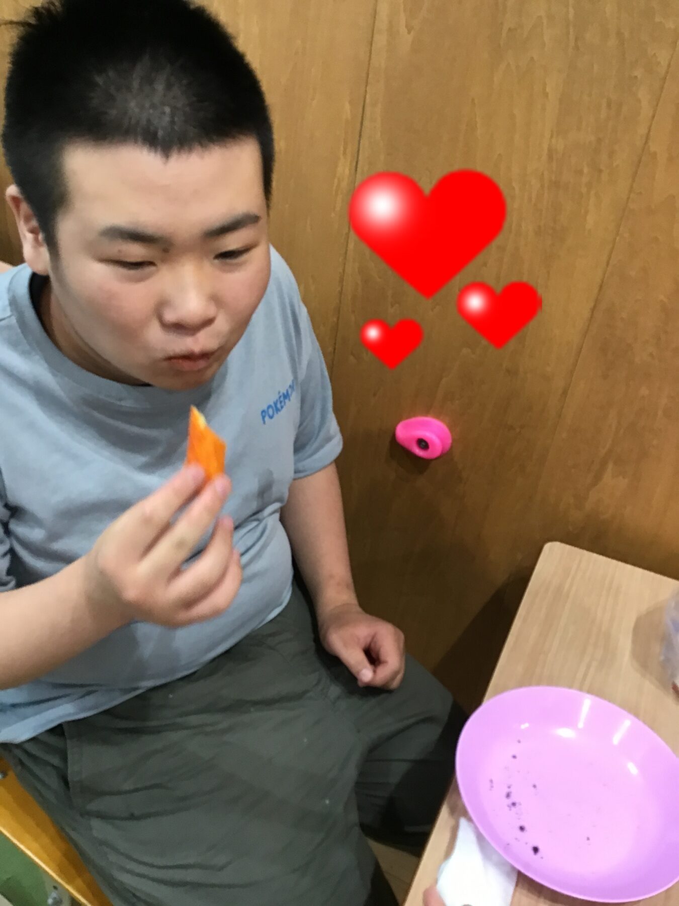 Young man in a gray shirt eating an orange snack at a table, with red heart emojis nearby.