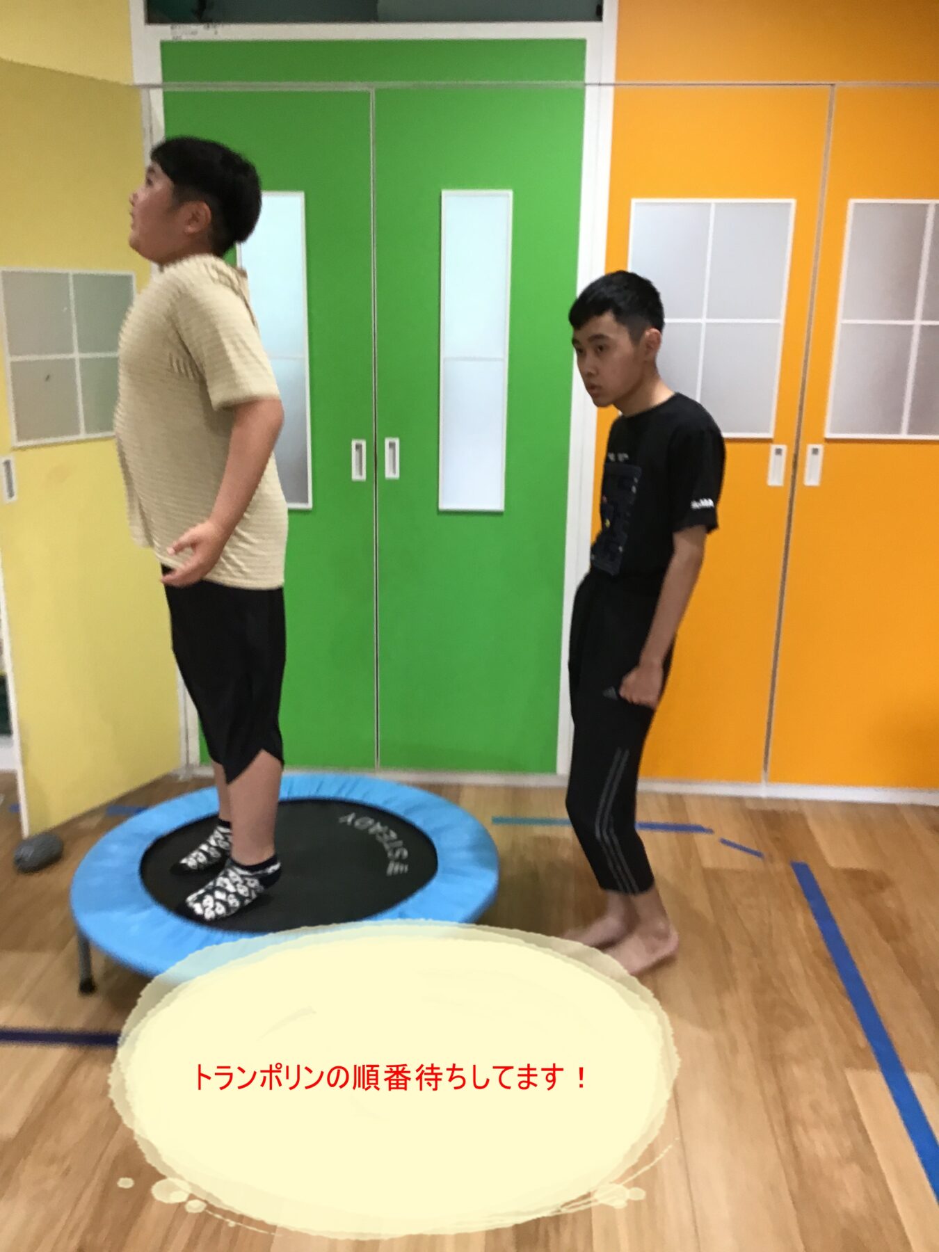 Two young men in a gym waiting on mini trampolines; one stands on a blue trampoline facing left, the other stands nearby near colorful green and orange lockers.