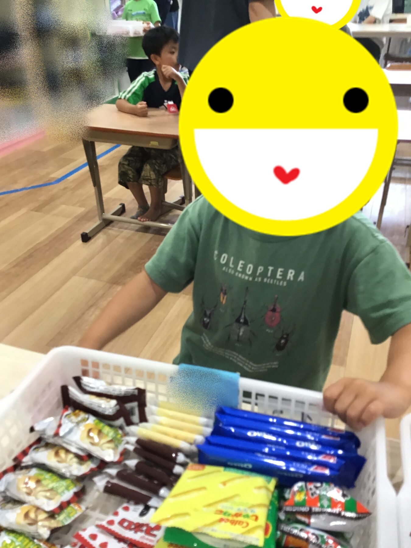 Child in a green shirt at a checkout with a basket of assorted snacks; a large yellow smiley-face graphic covers their face.