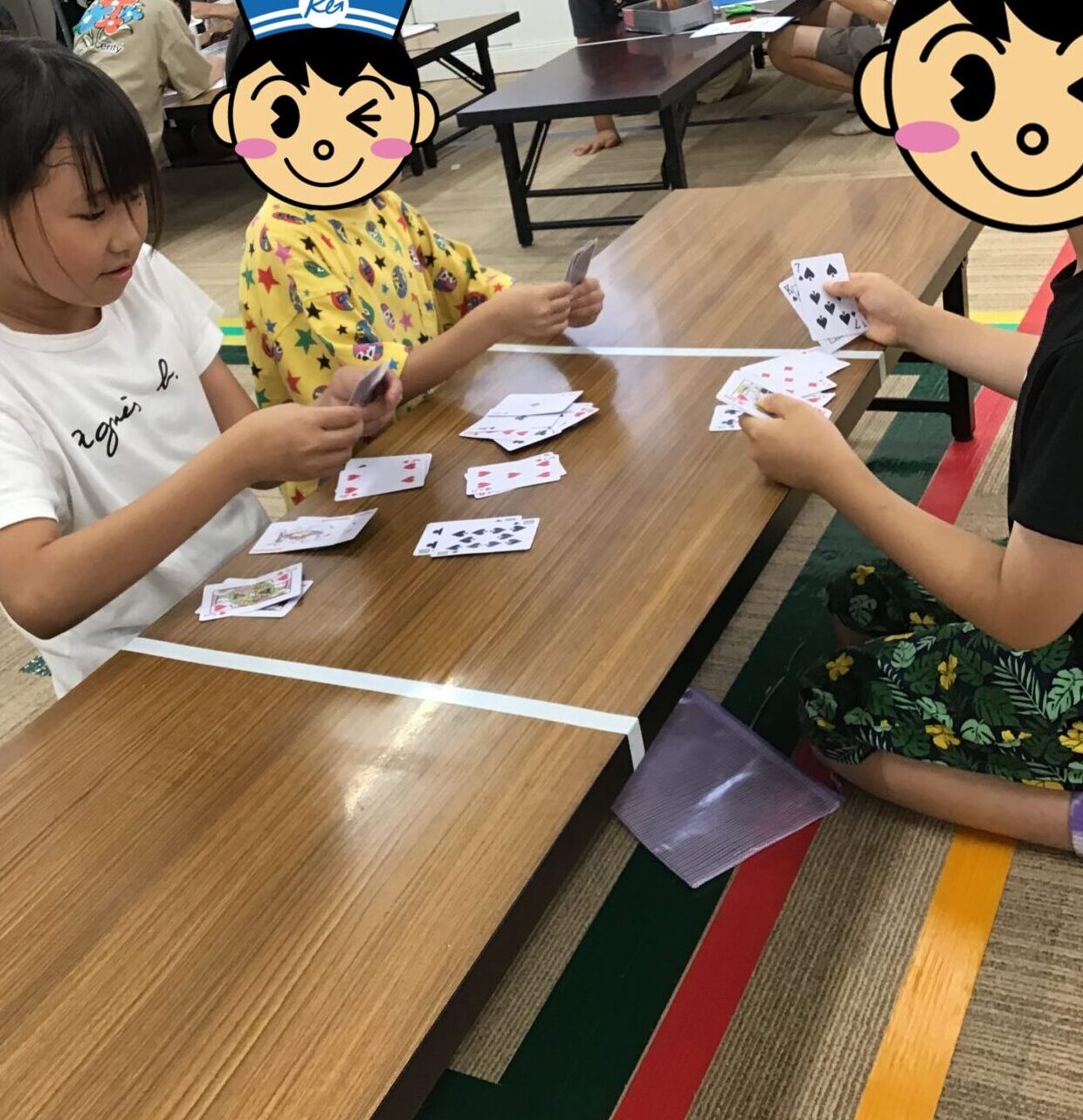 Children are seated at low wooden tables, playing cards together in a classroom setting as others work in the background