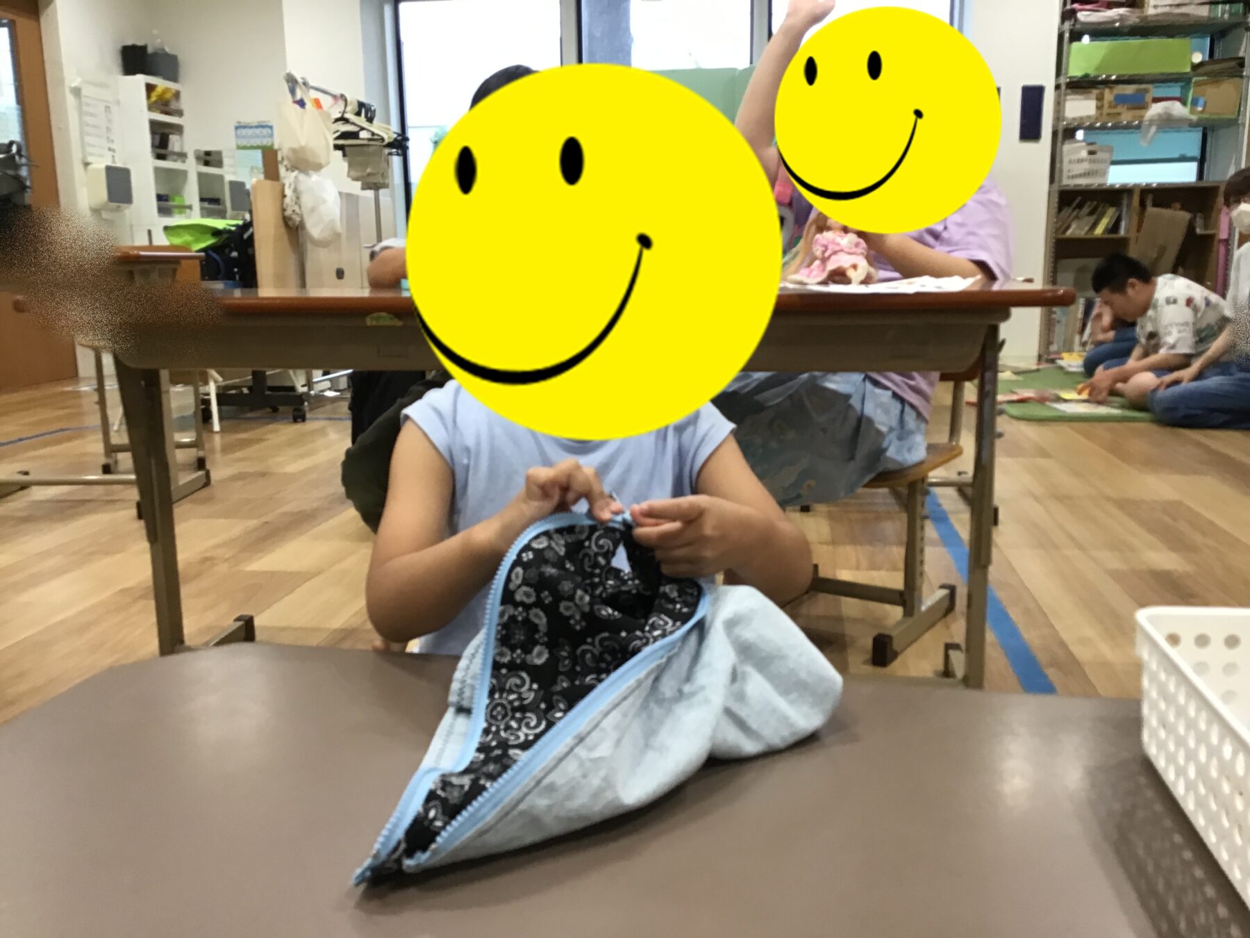 Children seated at a craft table in a classroom; the foreground child is opening a light blue backpack while two yellow smiley overlays obscure their faces, with shelves and supplies in the background.