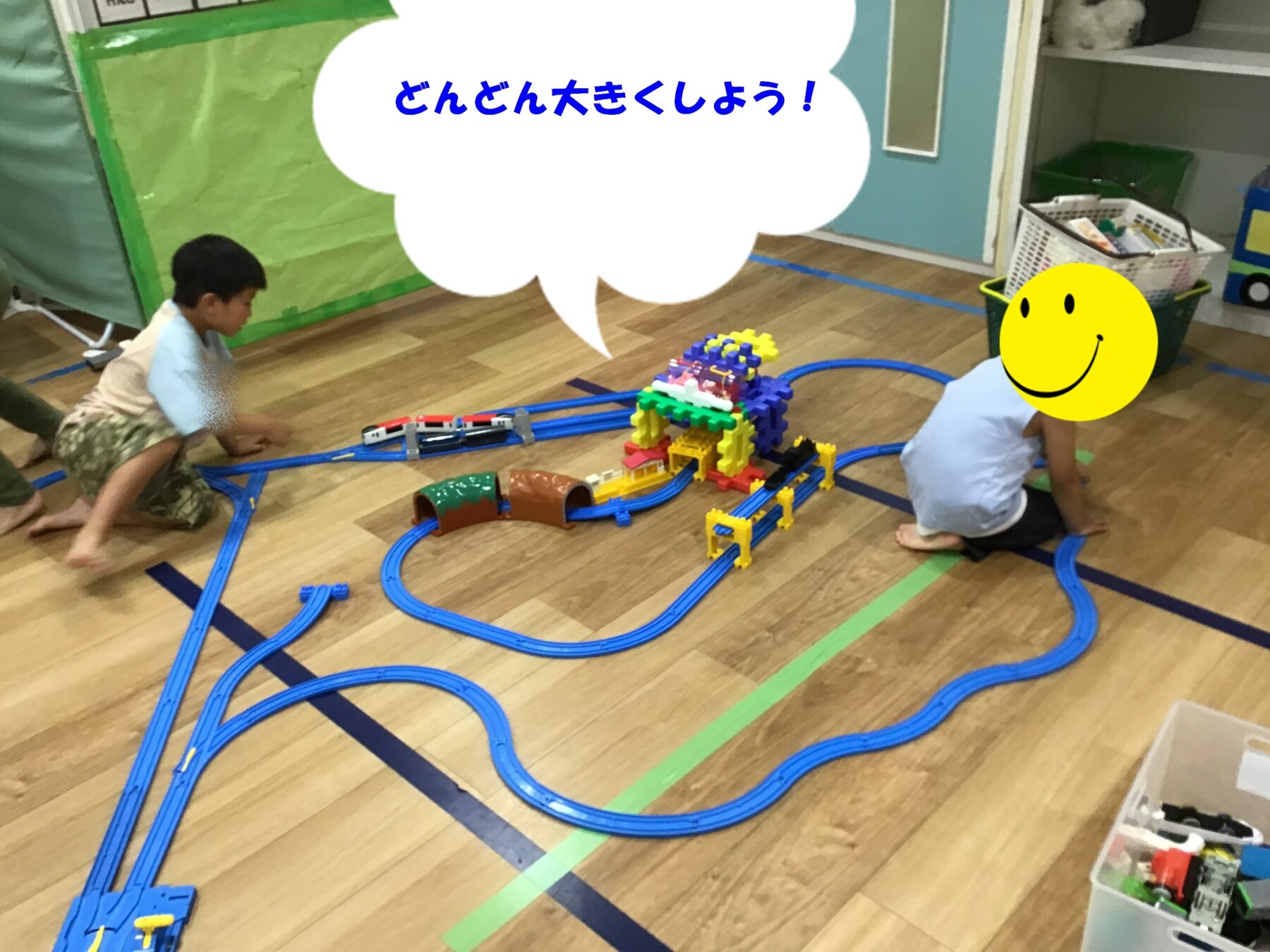Two children kneeling on a wooden floor build a blue toy train track layout with colorful plastic blocks and tunnels.