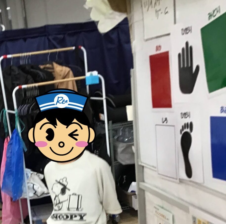 Child standing barefoot on a pink line in a classroom play area beside a clothing rack and wall charts with hand/foot icons.