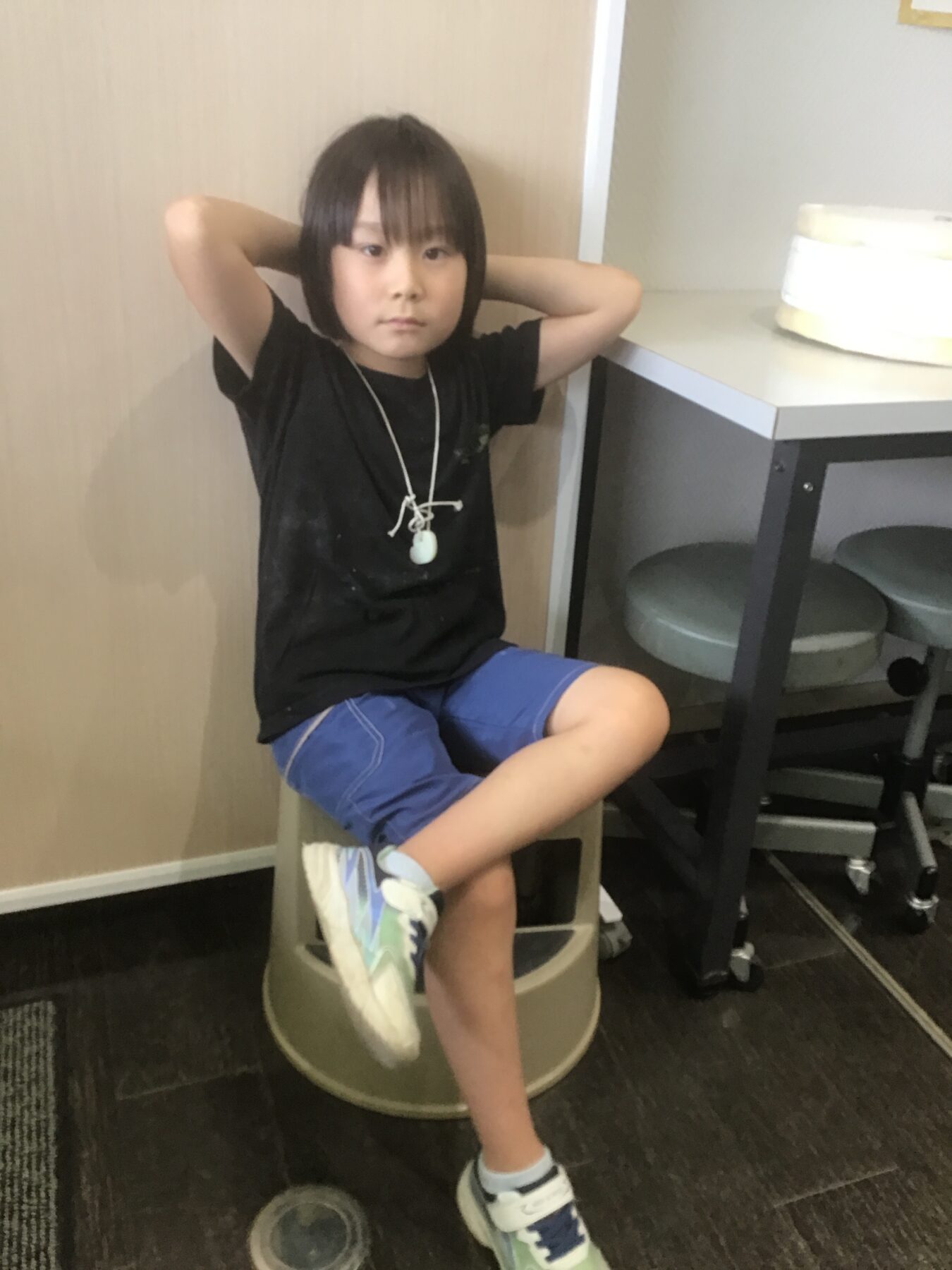 Young child sits on a stool with arms behind head in a casual pose, wearing a black shirt and blue shorts, indoors by a table with a cake nearby.