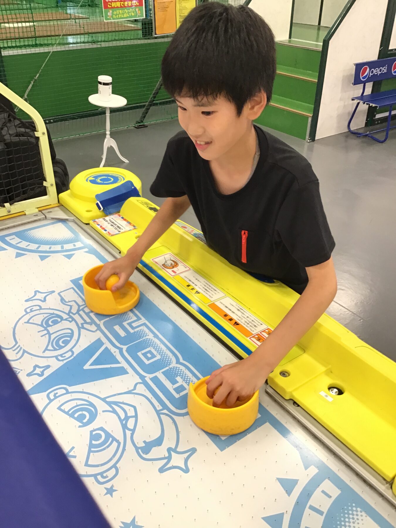 Boy in a black shirt grips yellow game handles at an arcade machine indoors, concentrating on the blue and yellow control panel.