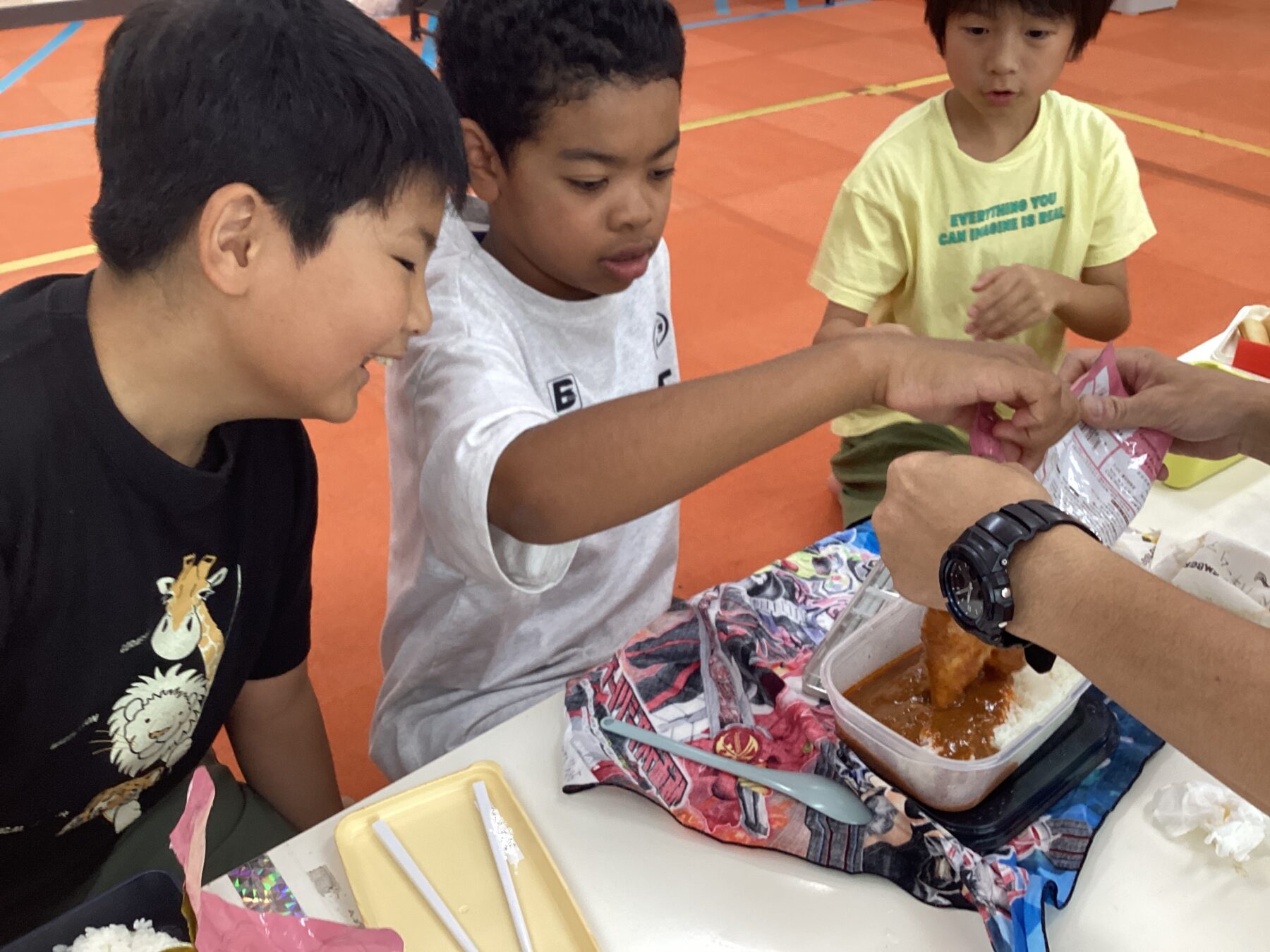 Three kids at a table share a curry meal; an adult hands over a pink packet to the group in a gym setting with an orange floor behind them.