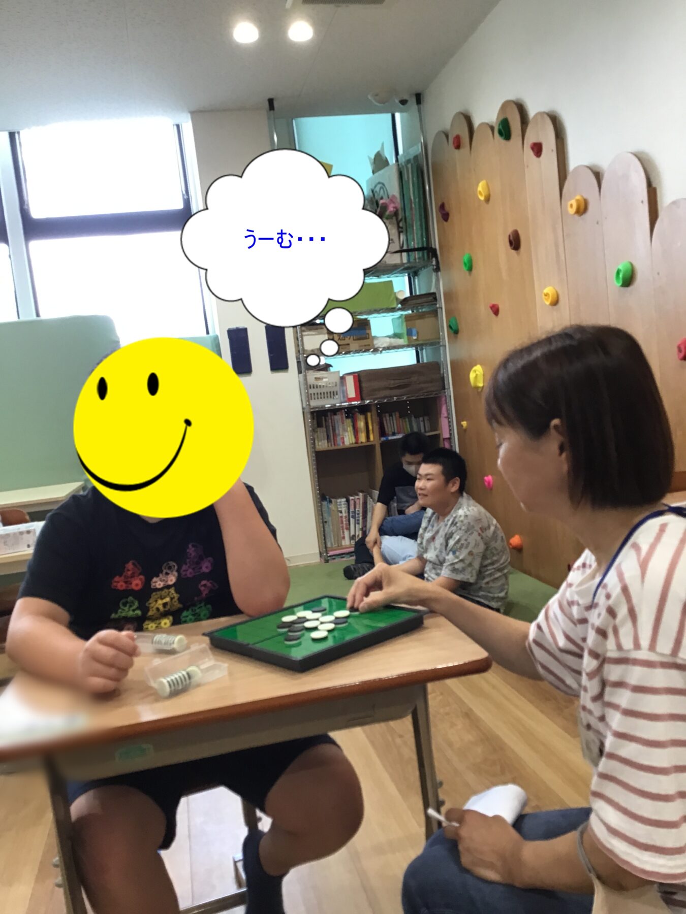 People sit around a table playing a board game with chips; a person has a large smiley-face overlay and a thought bubble reading うーむ… in a classroom with climbing wall panels in the background.