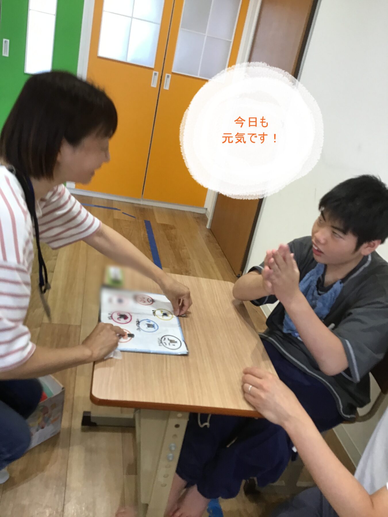 Woman and a man in a wheelchair sit at a wooden table, the woman pointing to a game board as they share a moment indoors near orange doors.