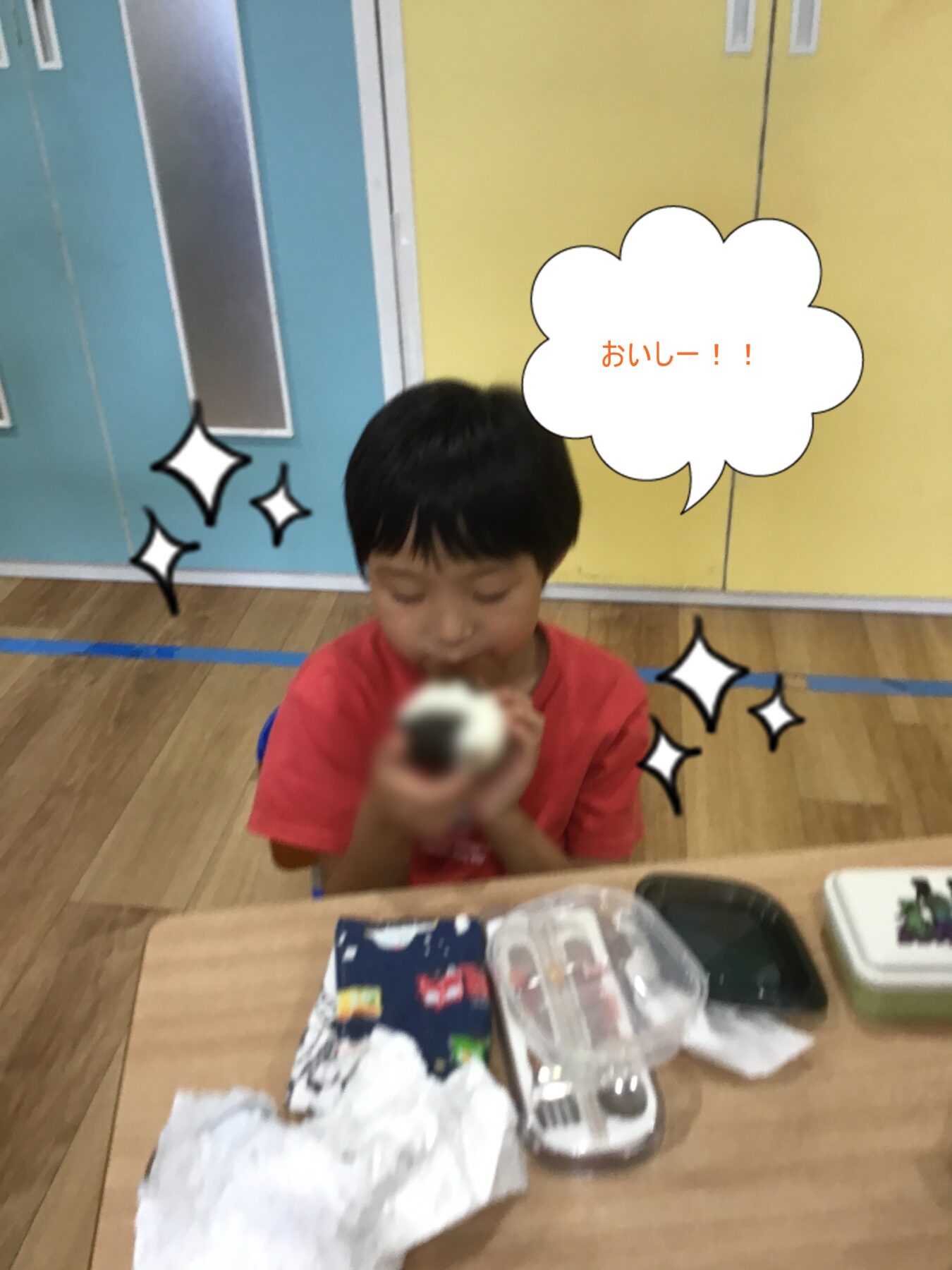 A young boy in a red shirt sits at a table in a classroom, holding food toward the camera with a drawn speech bubble saying “おいしー!!” (Delicious!), colorful lockers in the background.