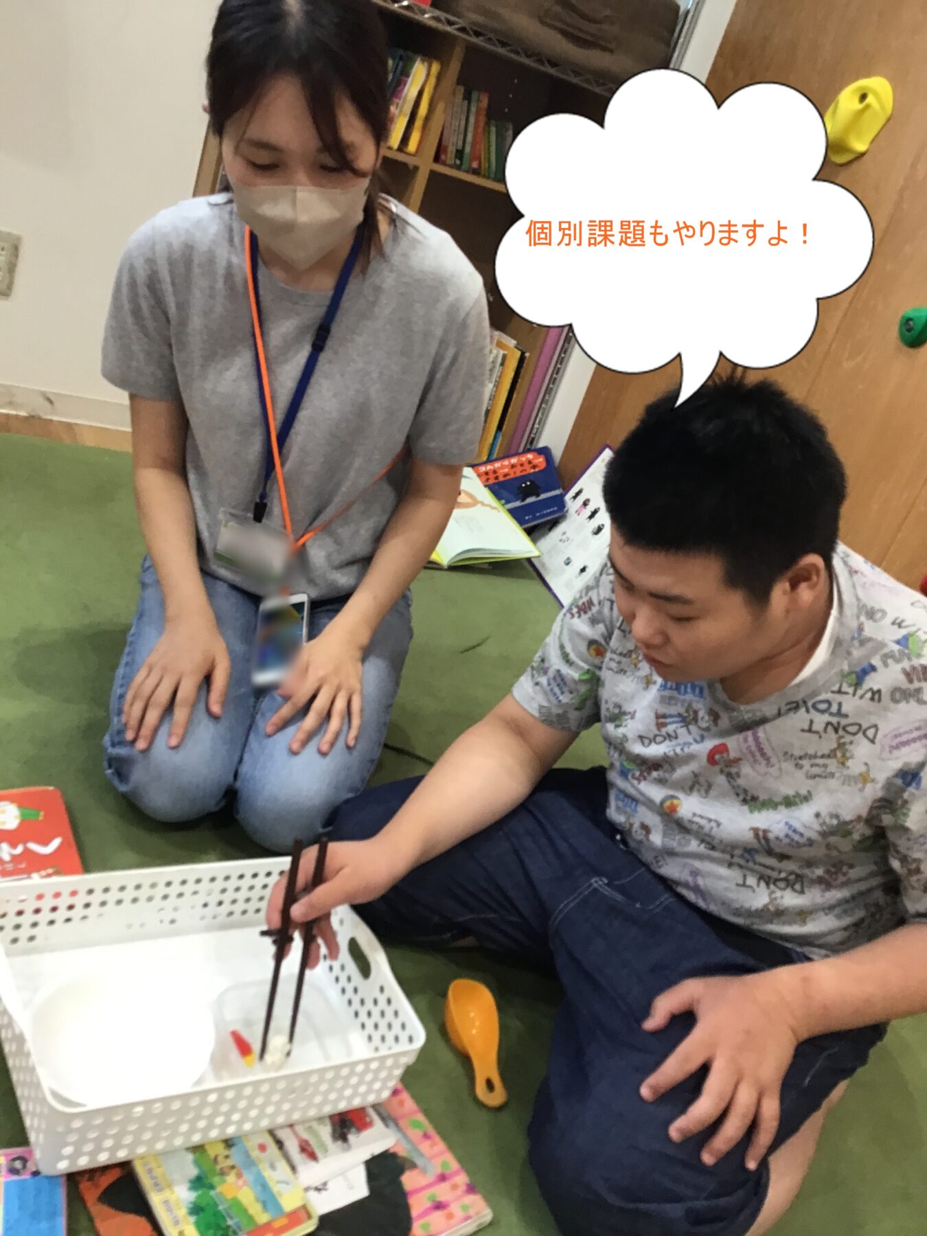 Woman in a gray shirt and mask kneels on a green mat beside a boy; they work on a hands-on activity with chopsticks over a white basket, books and colorful cards nearby.