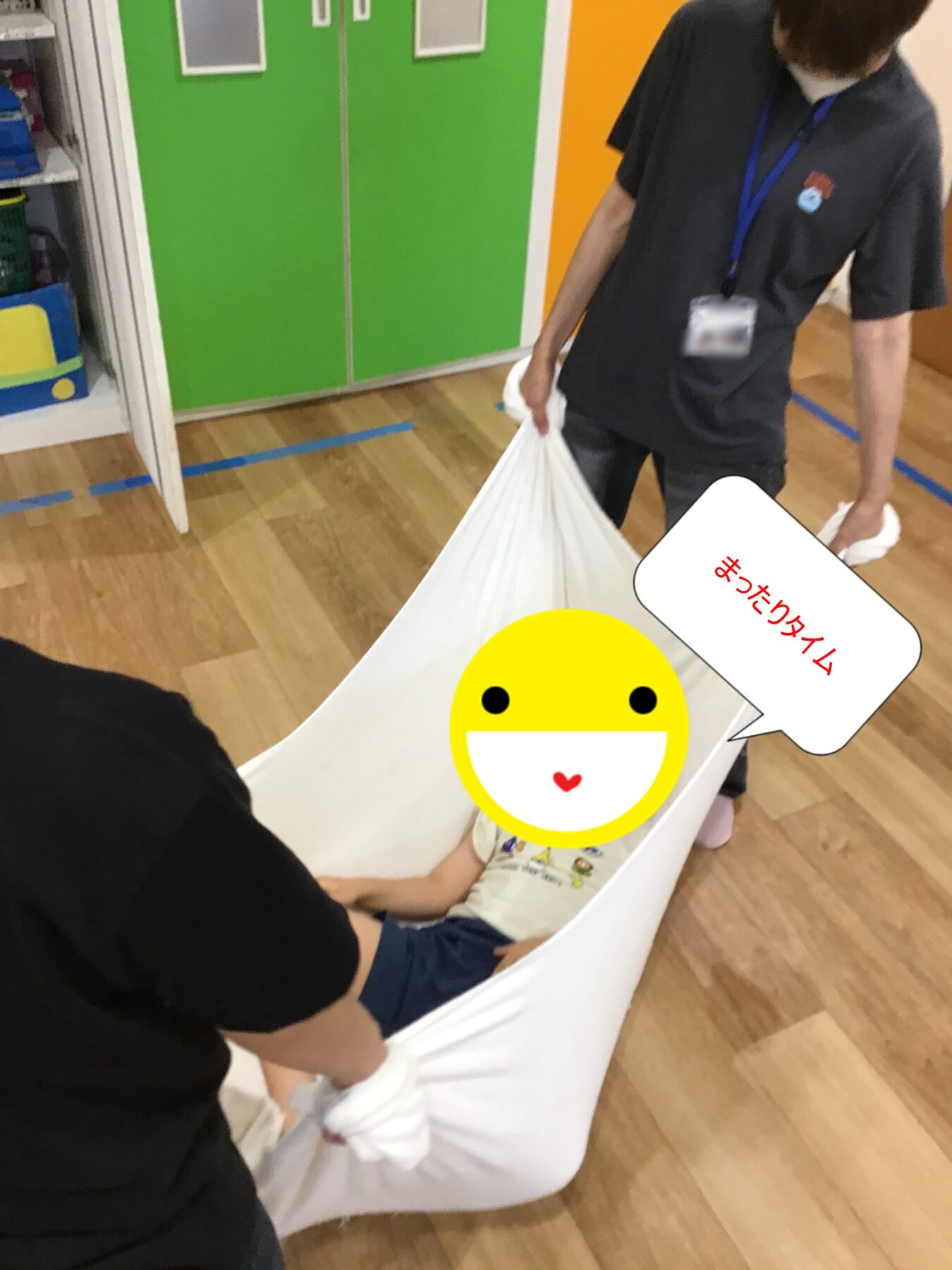 Adult in a gray shirt with a blue lanyard carries a large white fabric container holding a child, a big yellow smiley emoji covers part of the scene; background shows green cabinets and wooden floor; a speech bubble shows Japanese text まつりタイム (Festival time).