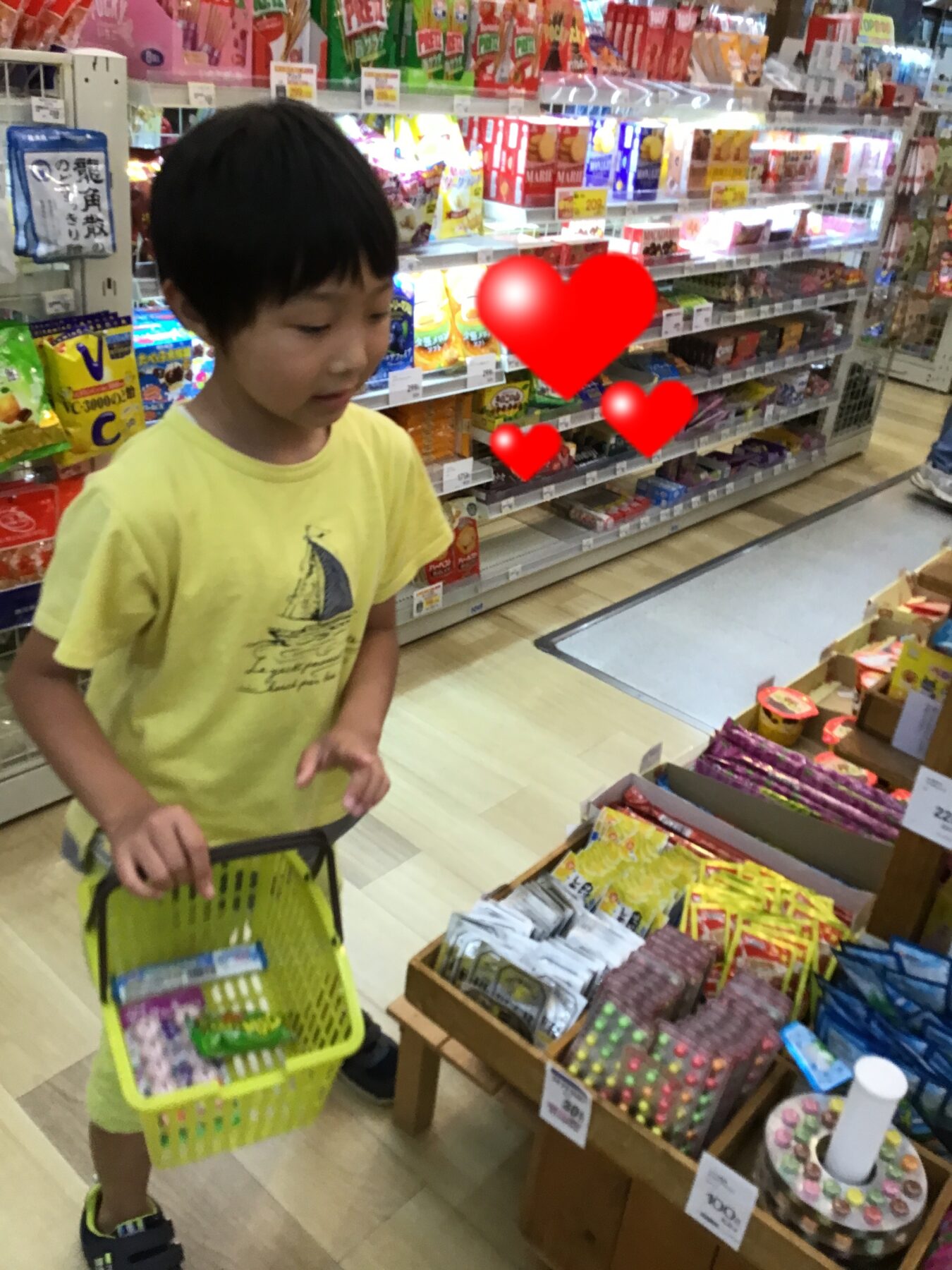 Young boy in a yellow shirt pushing a bright green shopping basket in a supermarket candy aisle with colorful snack shelves in the background.
