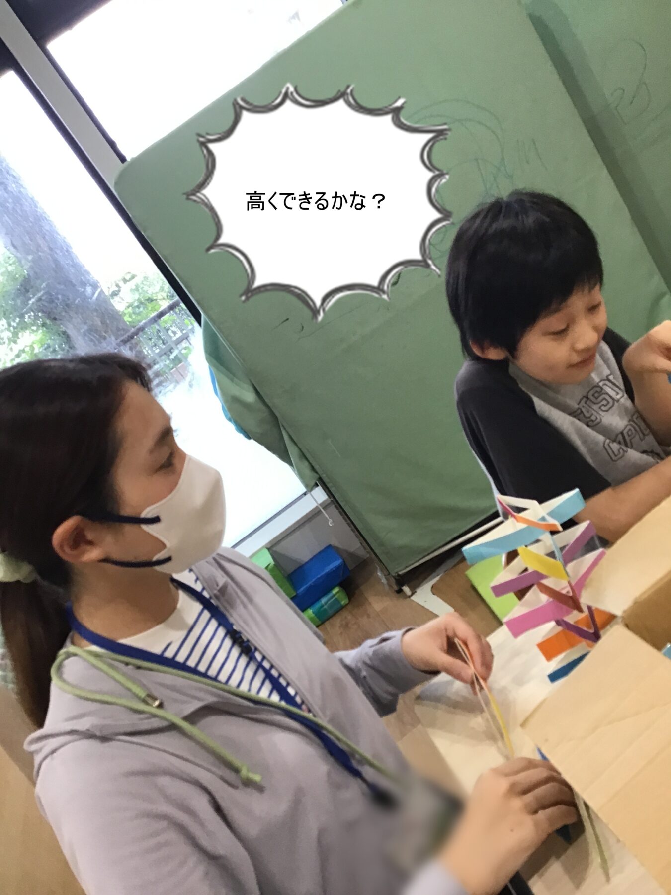 Two kids at a table making paper-chain crafts with colorful strips; speech bubble reads '高くできるかな?'