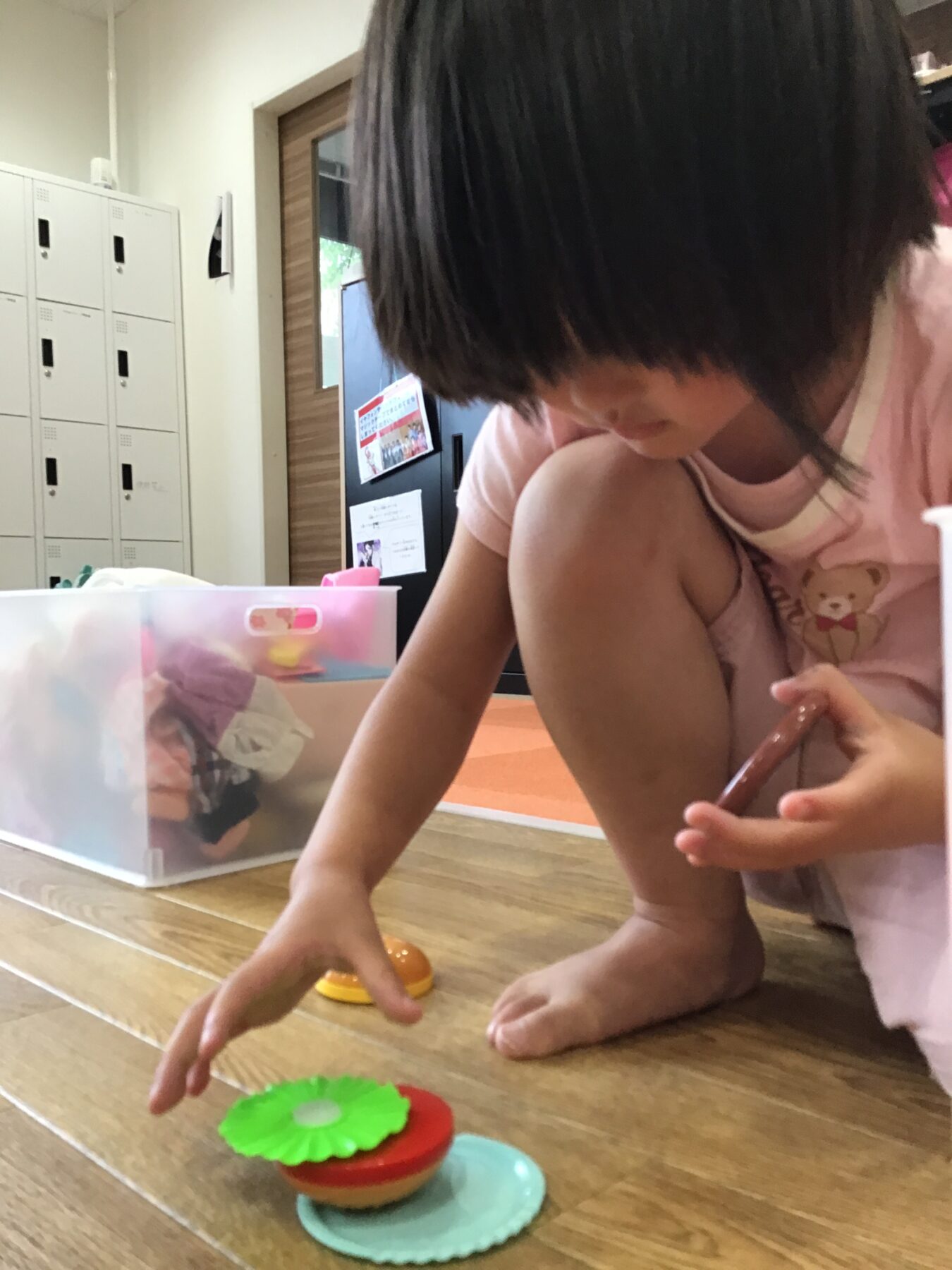 A young child kneels on a wooden floor, reaching for colorful toy lids stacked in front of her near a clear plastic bin in a classroom or playroom.