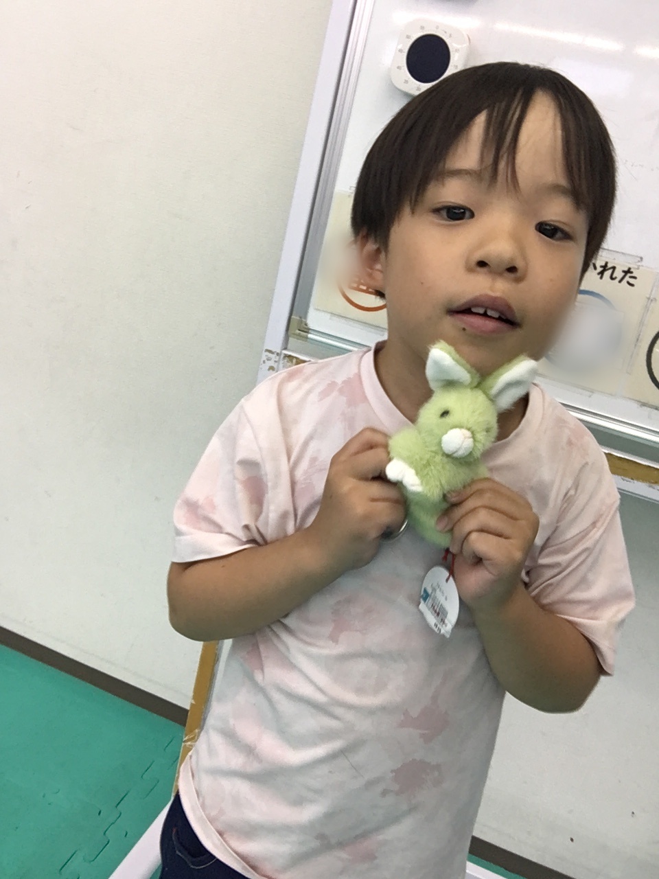 Young child in a pale pink shirt holding a small green plush bunny toy close to their chest indoors.
