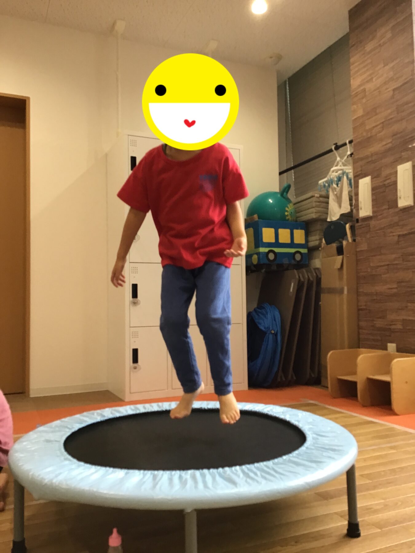 Child in red shirt and blue pants jumps on an indoor trampoline in a playroom, bare feet visible, with lockers and toy shelves in the background.