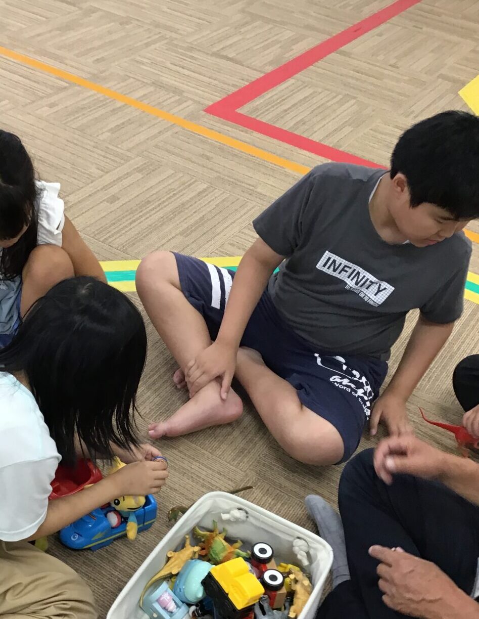 Children sit on a gym floor playing with assorted toy figures from a white tub nearby, colorful tape lines marking the floor.