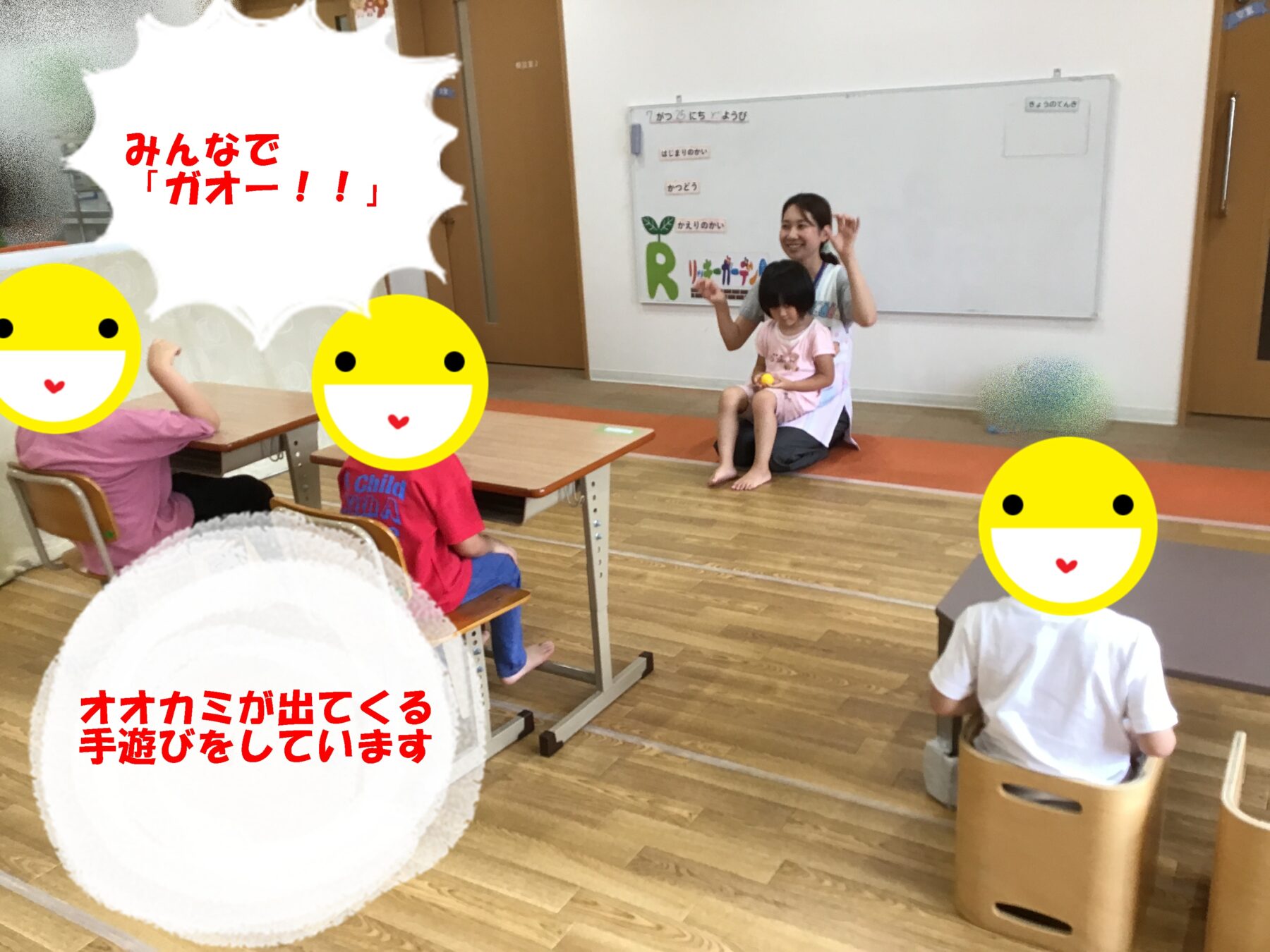 In a classroom, a teacher sits with a child on her lap while other kids at desks mimic actions; a large white speech bubble with red Japanese text 'みんなでガオー!!' dominates the left side, and yellow smiley stickers cover most faces. A whiteboard and classroom decor are in the background.