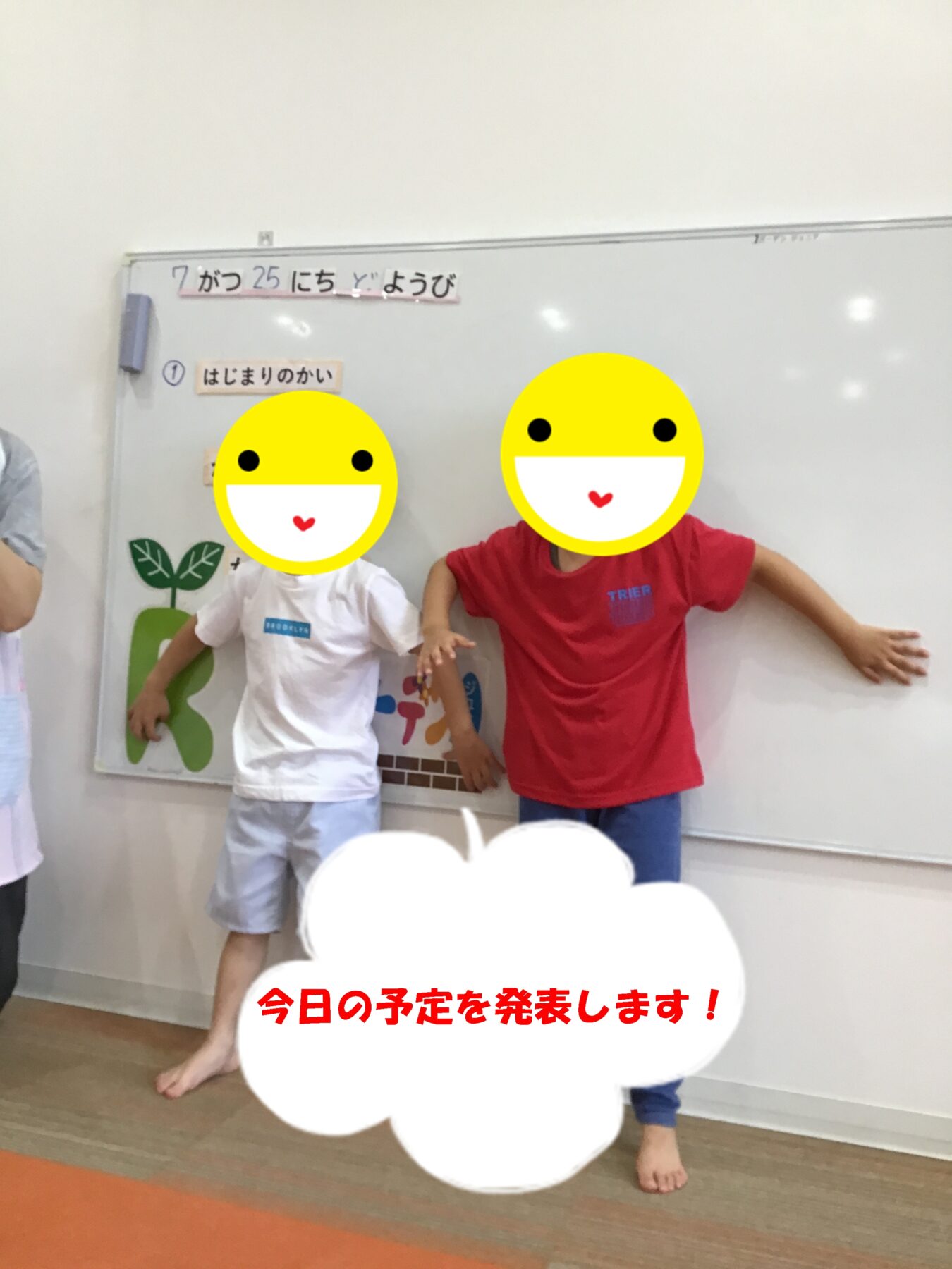 Two children pose in a classroom in front of a whiteboard, with large yellow smiley overlays covering their faces and red Japanese caption at the bottom.