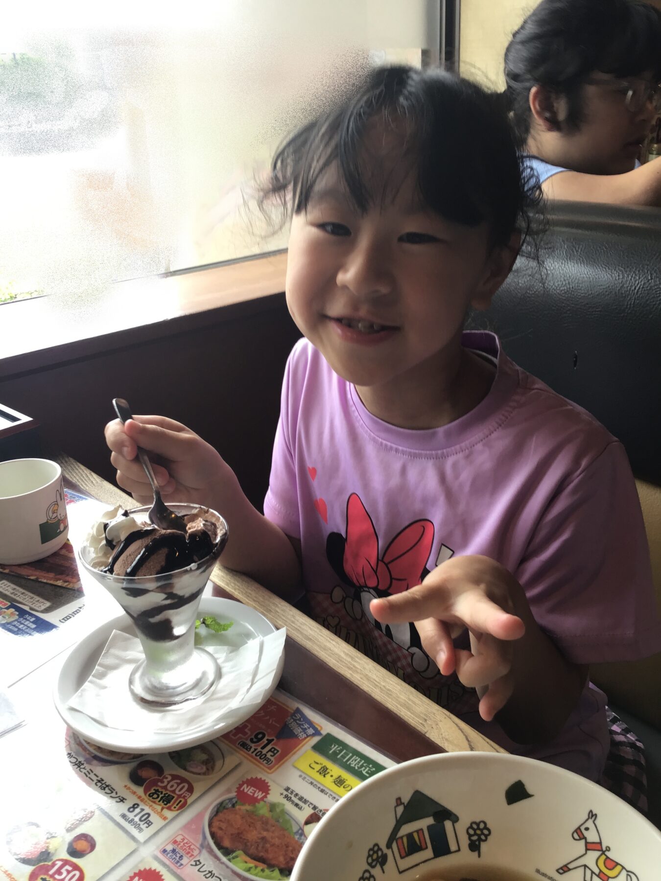 Smiling young girl in a pink shirt with a chocolate sundae, spoon in hand, at a cafe table.
