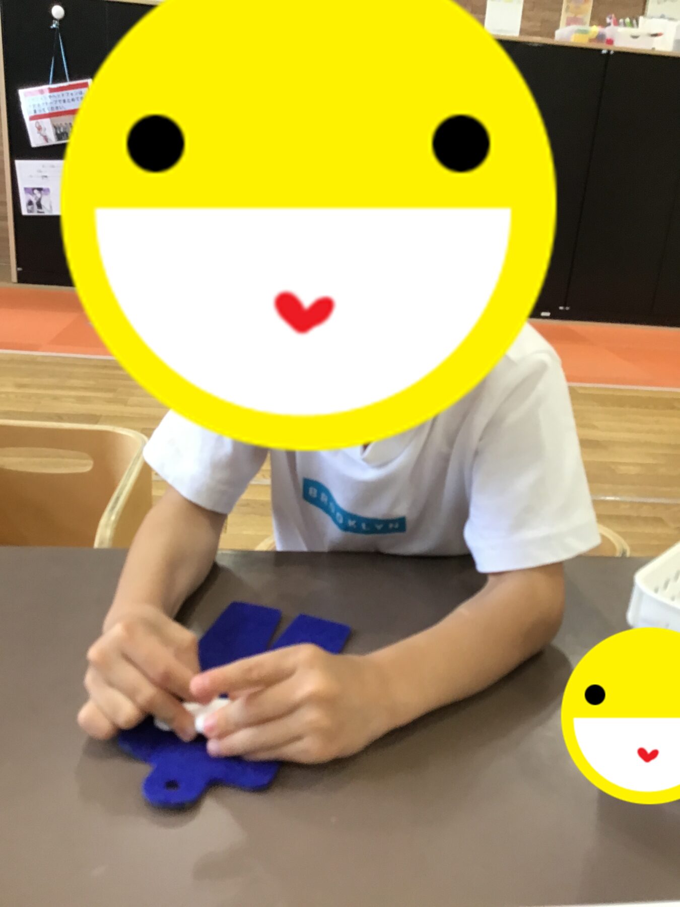 Child seated at a classroom table, arranging blue foam pieces, with a large yellow smiley graphic covering their face.
