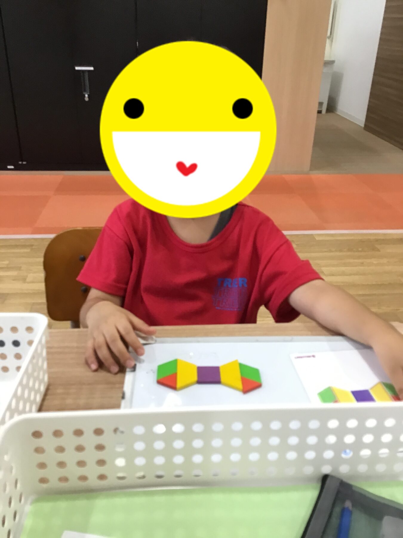 Child in a red shirt sits at a desk, assembling colorful tangram puzzle pieces on a white tray in a classroom setting.