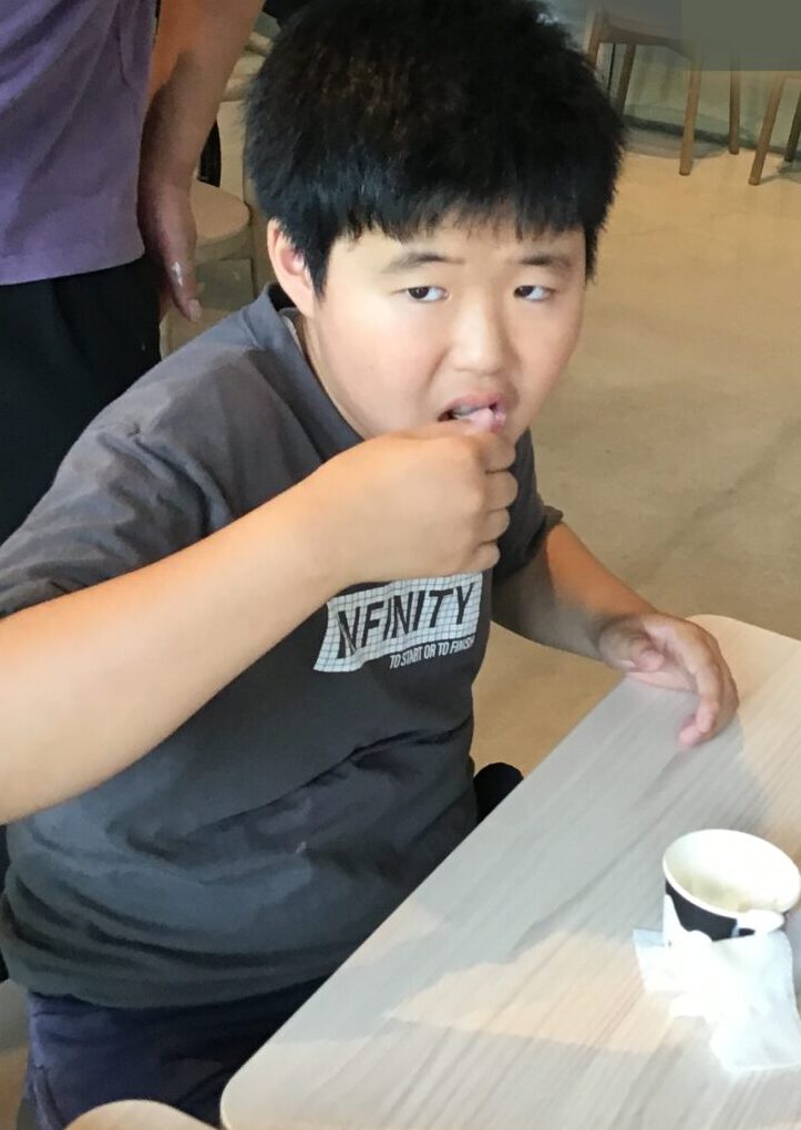 Young boy with dark hair eating at a light wood table in a casual dining area; a girl sits at a nearby table in a brown dress in the background.