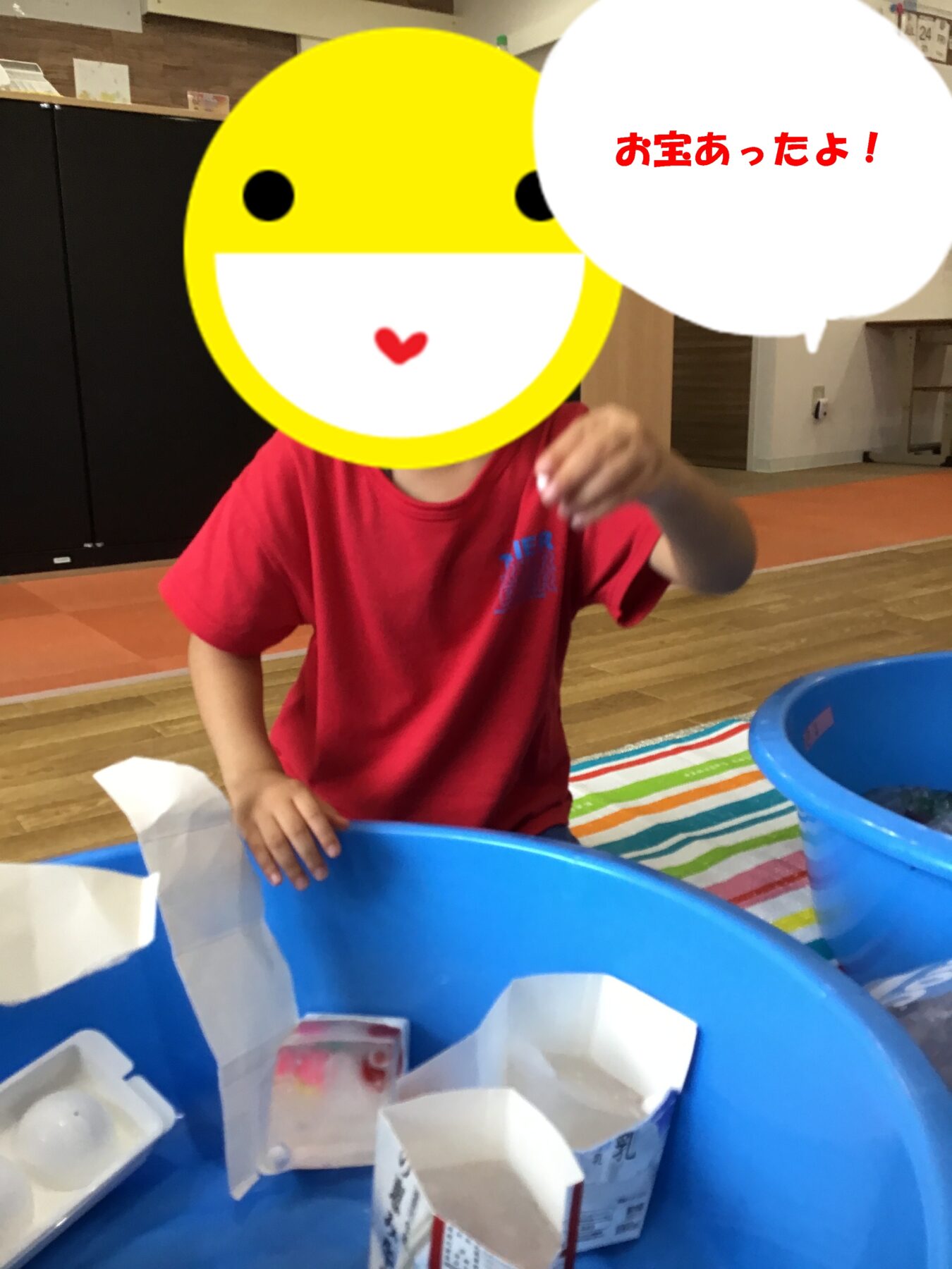 Child with a yellow smiley face mask sitting at a blue tub, reaching for paper treasures; speech bubble in Japanese says, 'I found treasure!'
