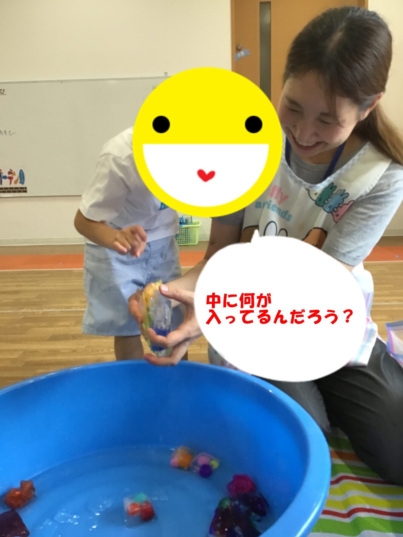 Child helping another child pick colorful toy pieces from a blue tub of water; a large yellow smiley face covers part of the scene.