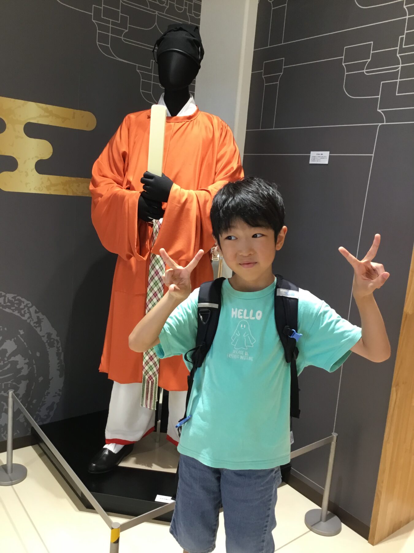 Young boy posing beside a life-size statue of a monk in orange robes, making peace signs with both hands.