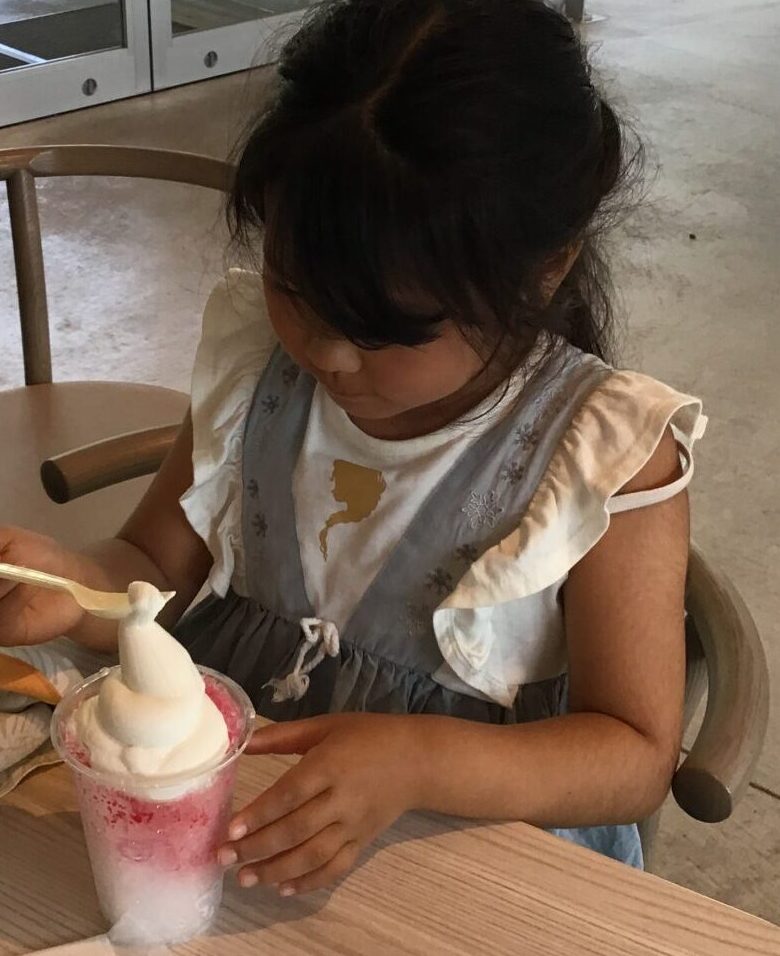 Young girl sits at a cafe table, eating dessert with a spoon while a pink-and-white whipped-cream drink sits in front of her on the table outside a glass-walled cafe area.